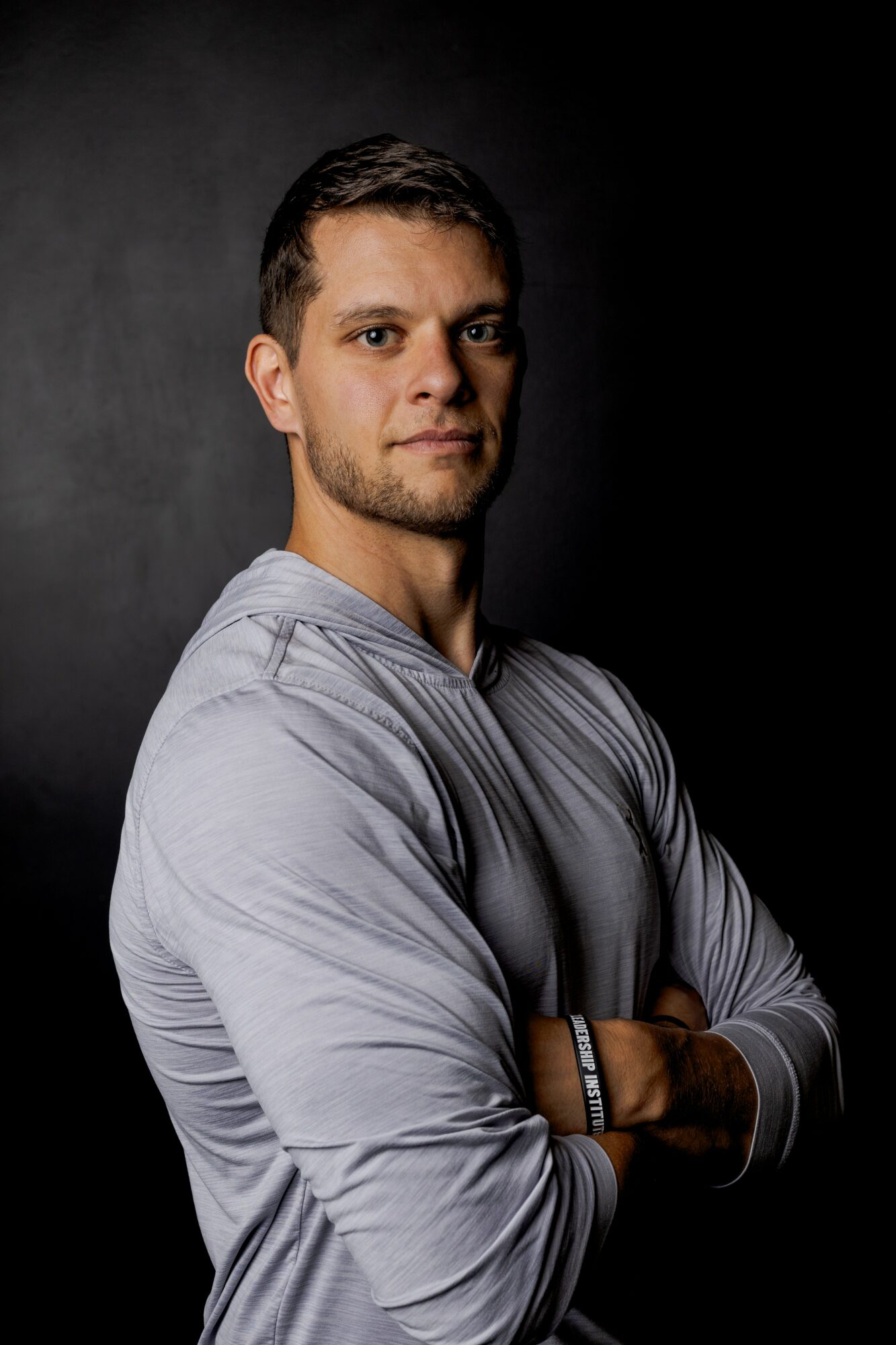 Young man with short dark hair and light skin, wearing a gray long-sleeve shirt, standing with arms crossed against a dark background.