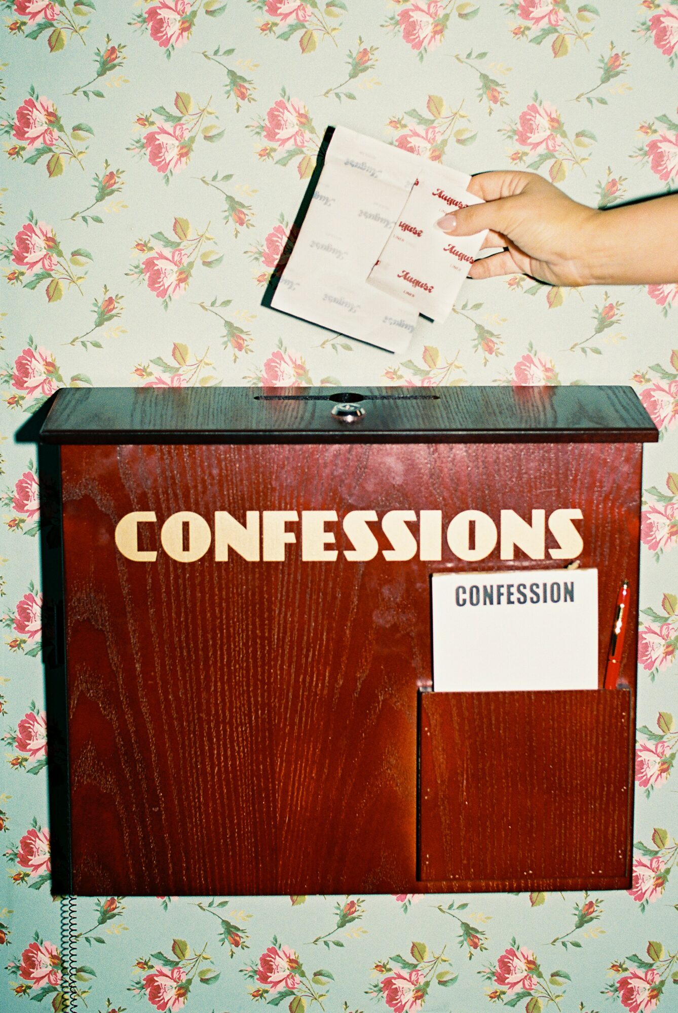 Hand placing paper into a wooden confession box against floral wallpaper background.