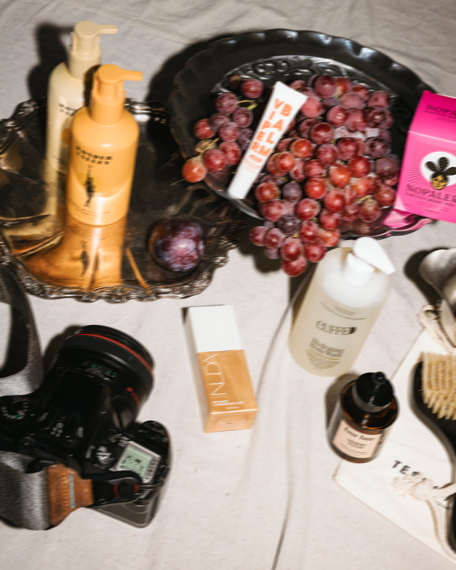 Black tray with grapes, skincare bottles, and a pink box, on a light surface with a camera and other items nearby.