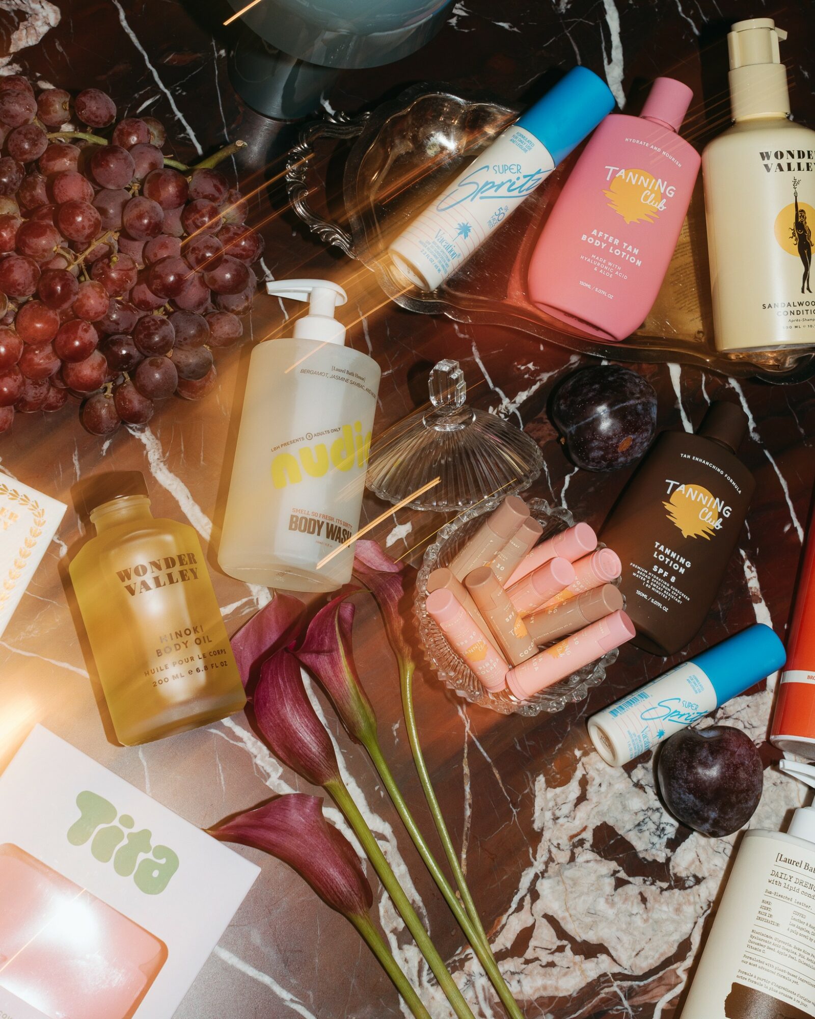 Flat lay of skincare, haircare products, grapes, cherries, and flowers on a marble surface.