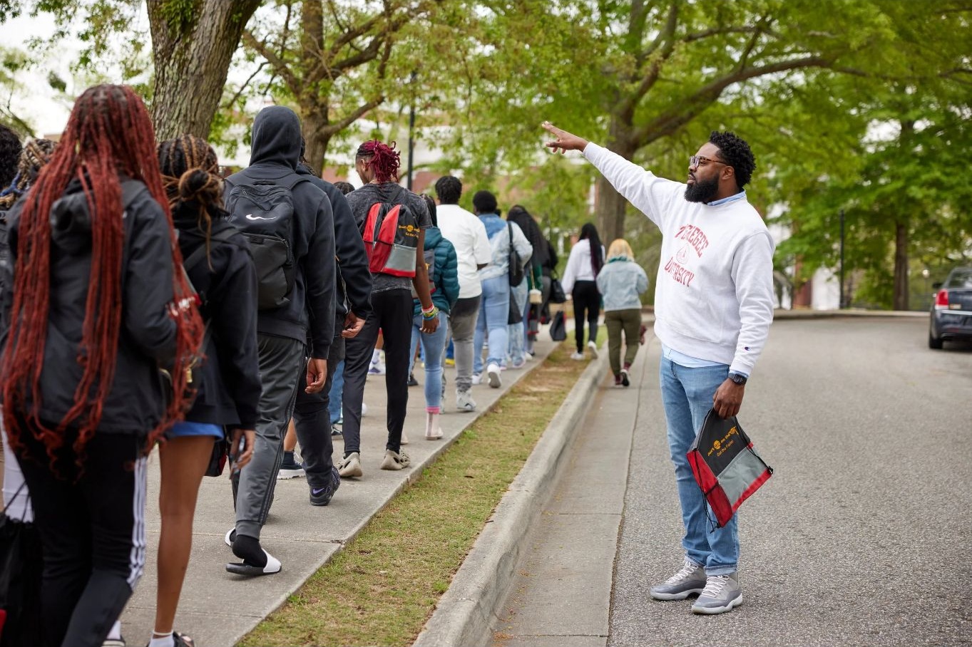 Man guides a line of people outdoors on a sidewalk, trees in background, holding a clipboard, gesturing with his right hand.