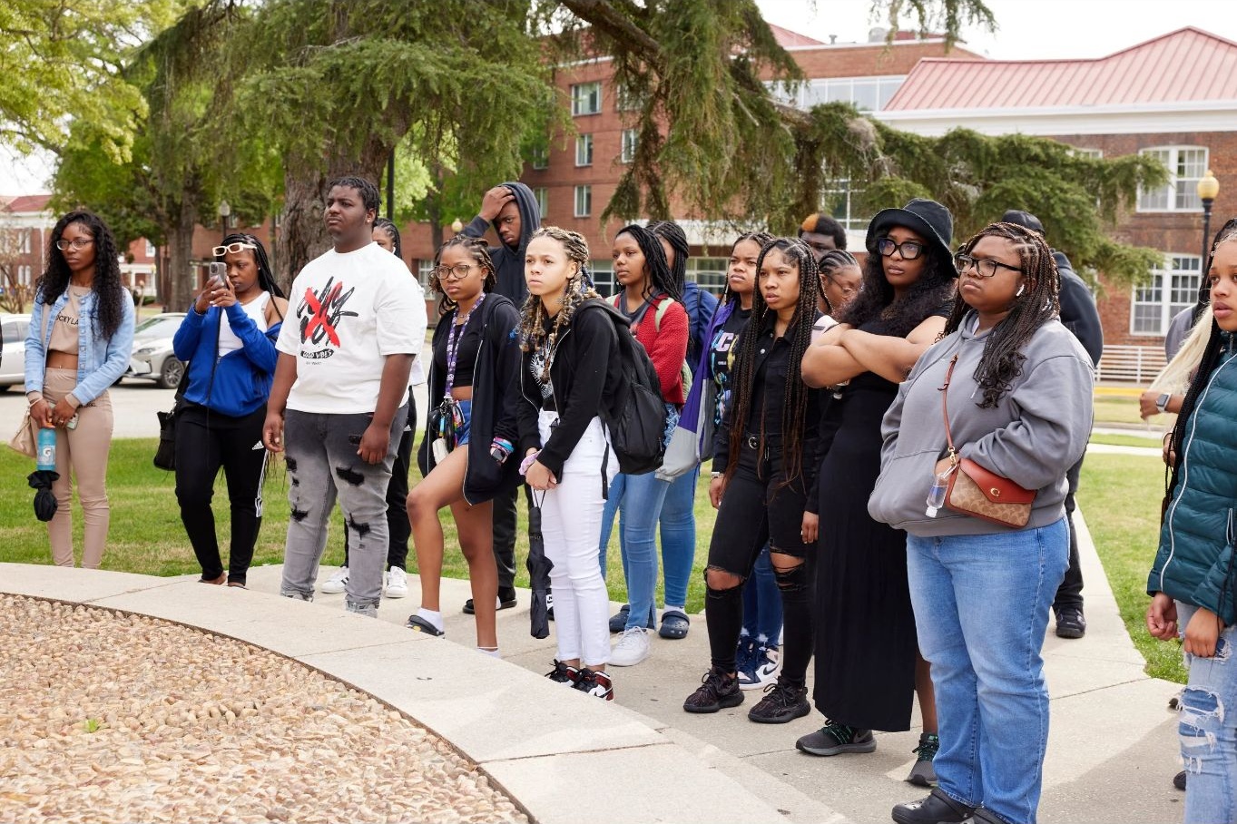Group of young people standing outdoors on a sidewalk, some with backpacks, in front of trees and buildings.