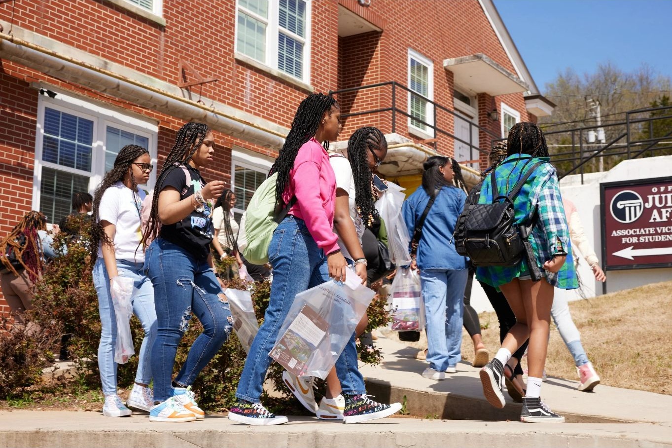 Group of young people walking outside a brick building, some carrying backpacks and bags, on a sunny day.