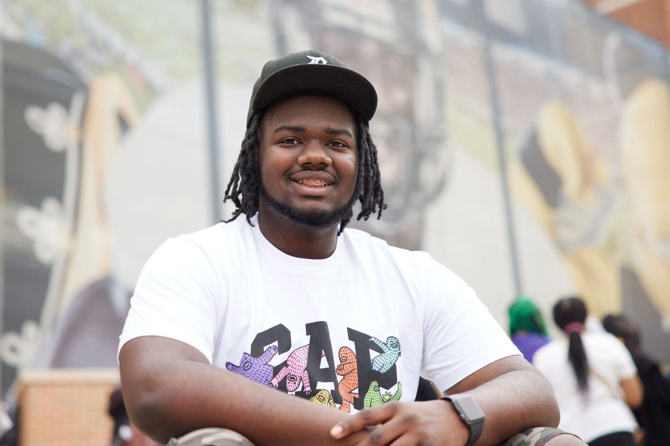 Smiling man with dreadlocks wearing a white T-shirt and black cap, sitting outdoors with a blurred background.