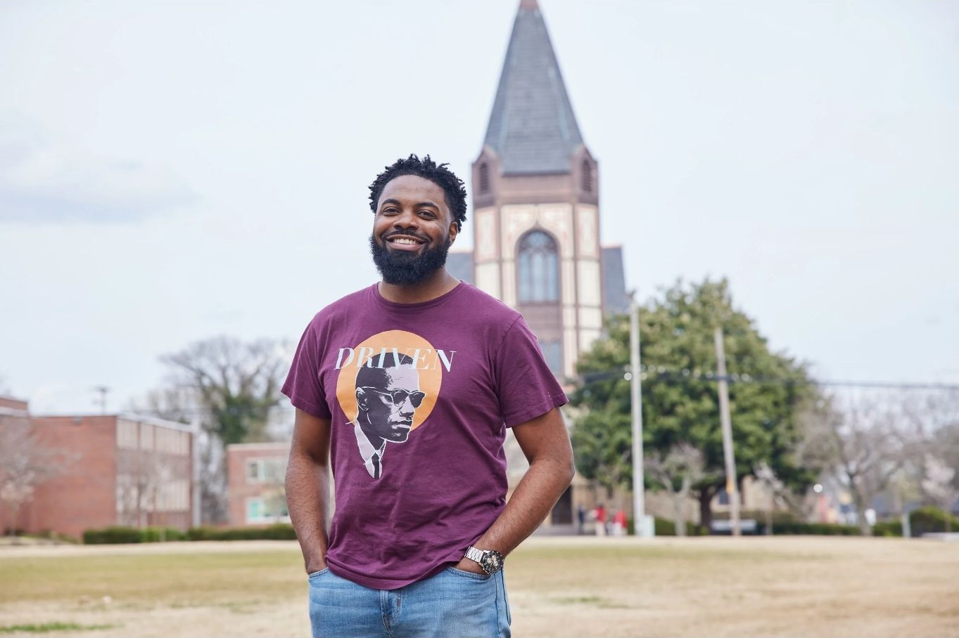 Man with a beard smiling outdoors, wearing a purple T-shirt with a graphic, in front of a church and trees.