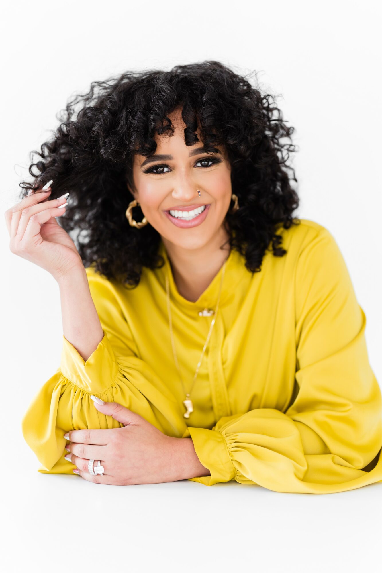 Woman with curly hair smiling, wearing a yellow blouse, gold hoop earrings, and a ring, against a white background.