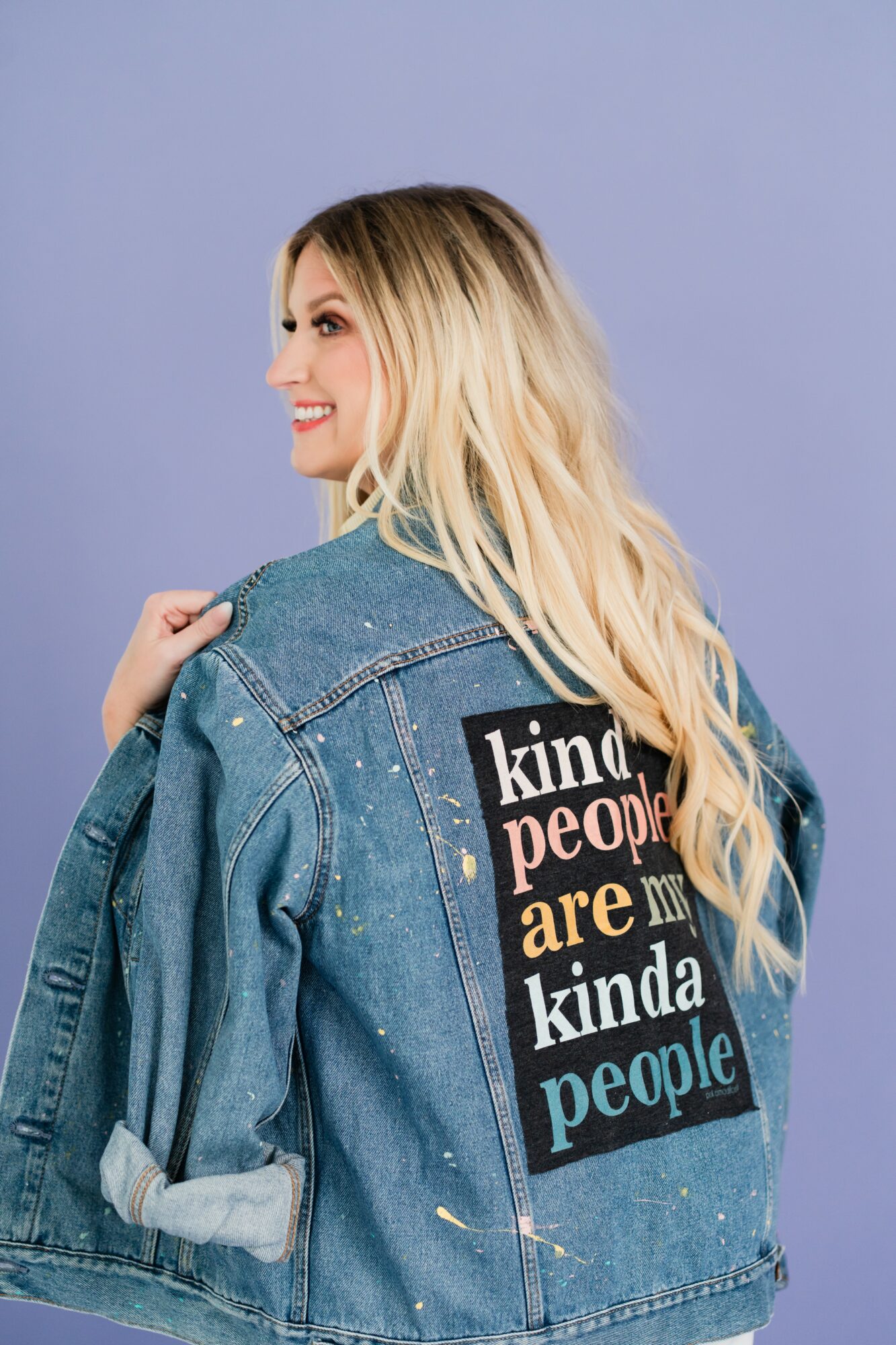 Woman with long blonde hair wearing a denim jacket with a colorful patch on the back, smiling, against a light purple background.