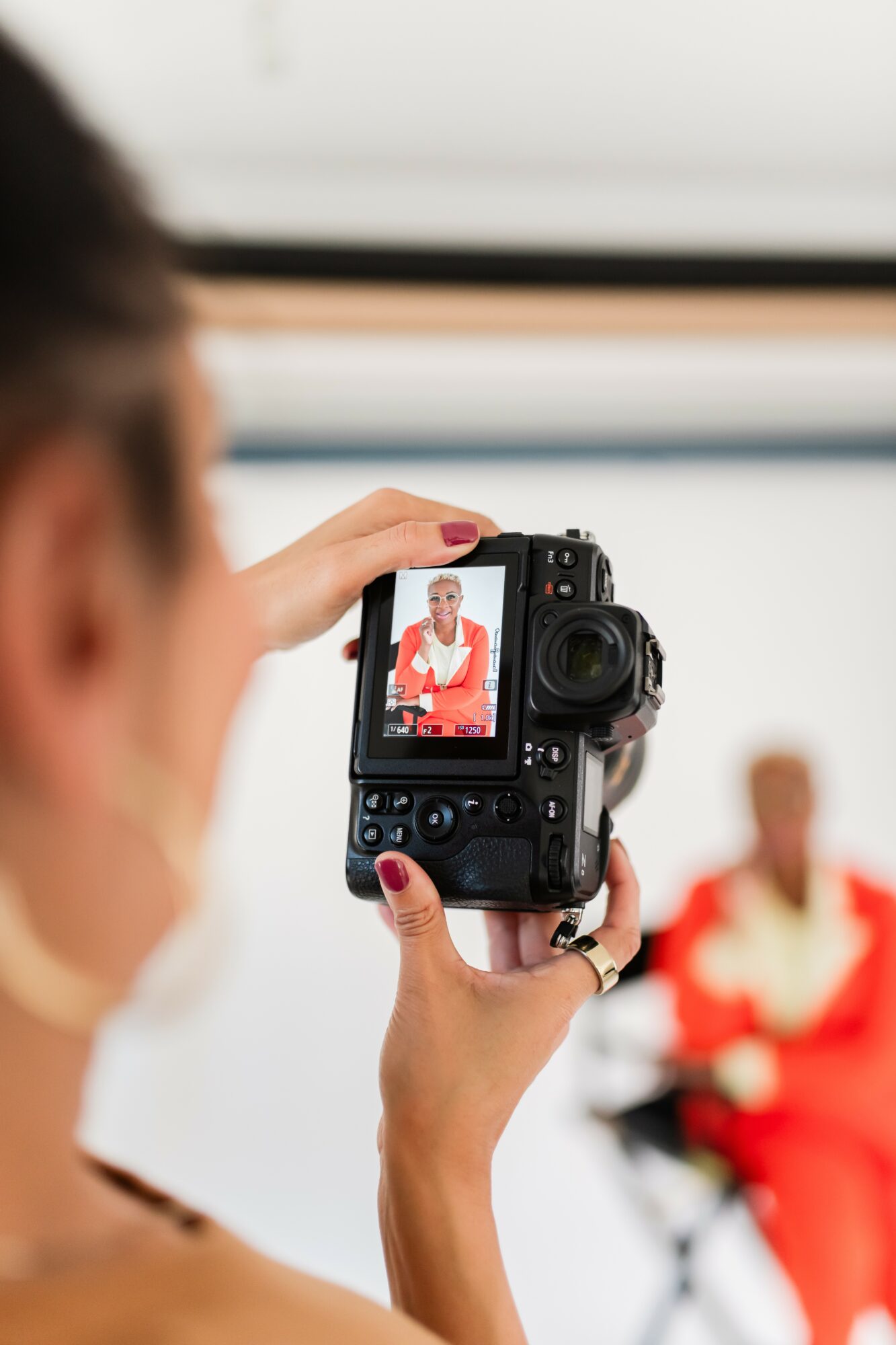 Person taking a photo of a woman in a red outfit with a digital camera, indoors, with a blurred background.