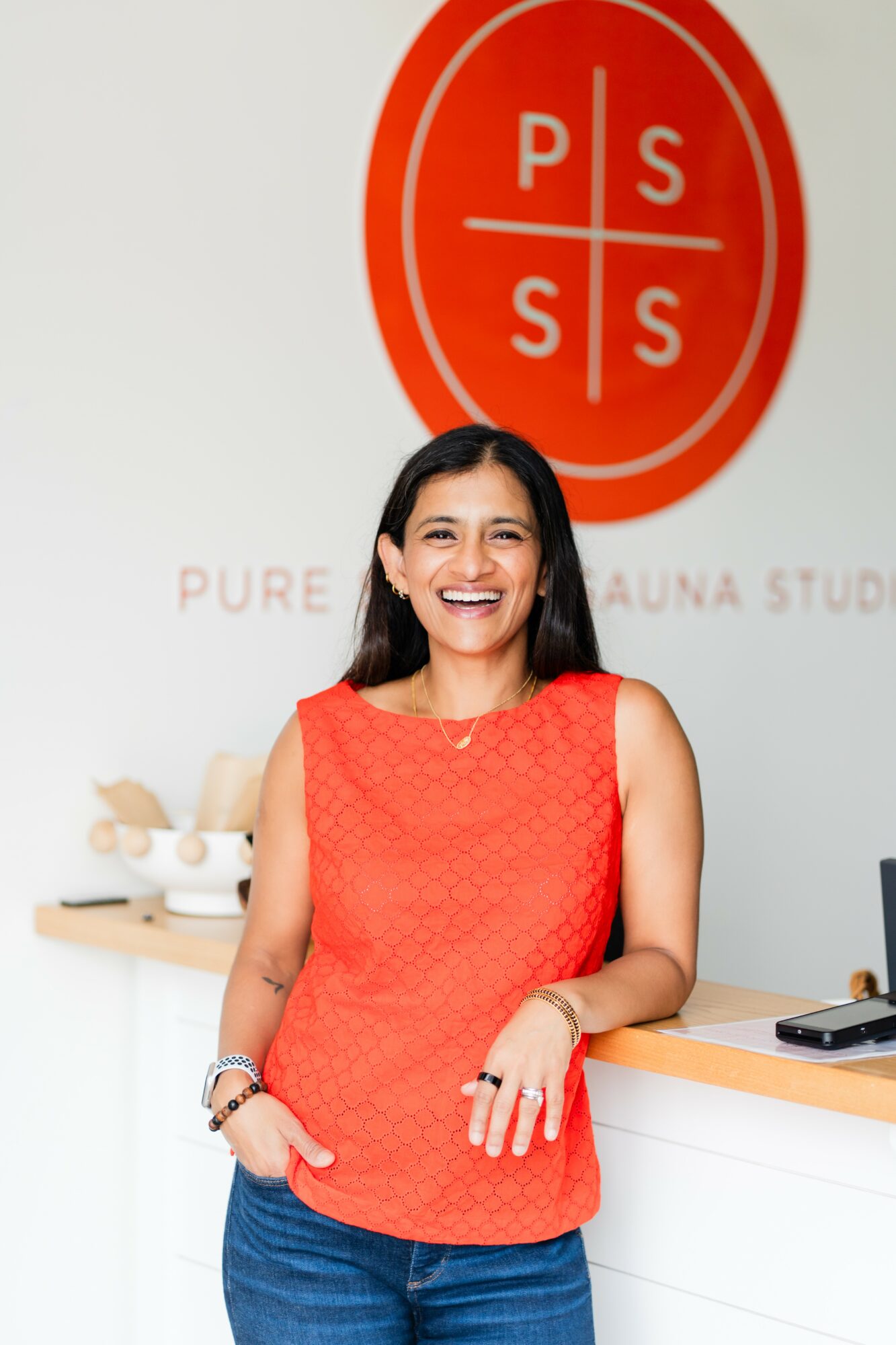 Smiling woman in red sleeveless top and jeans standing in front of a spa logo, with a counter and spa items nearby.