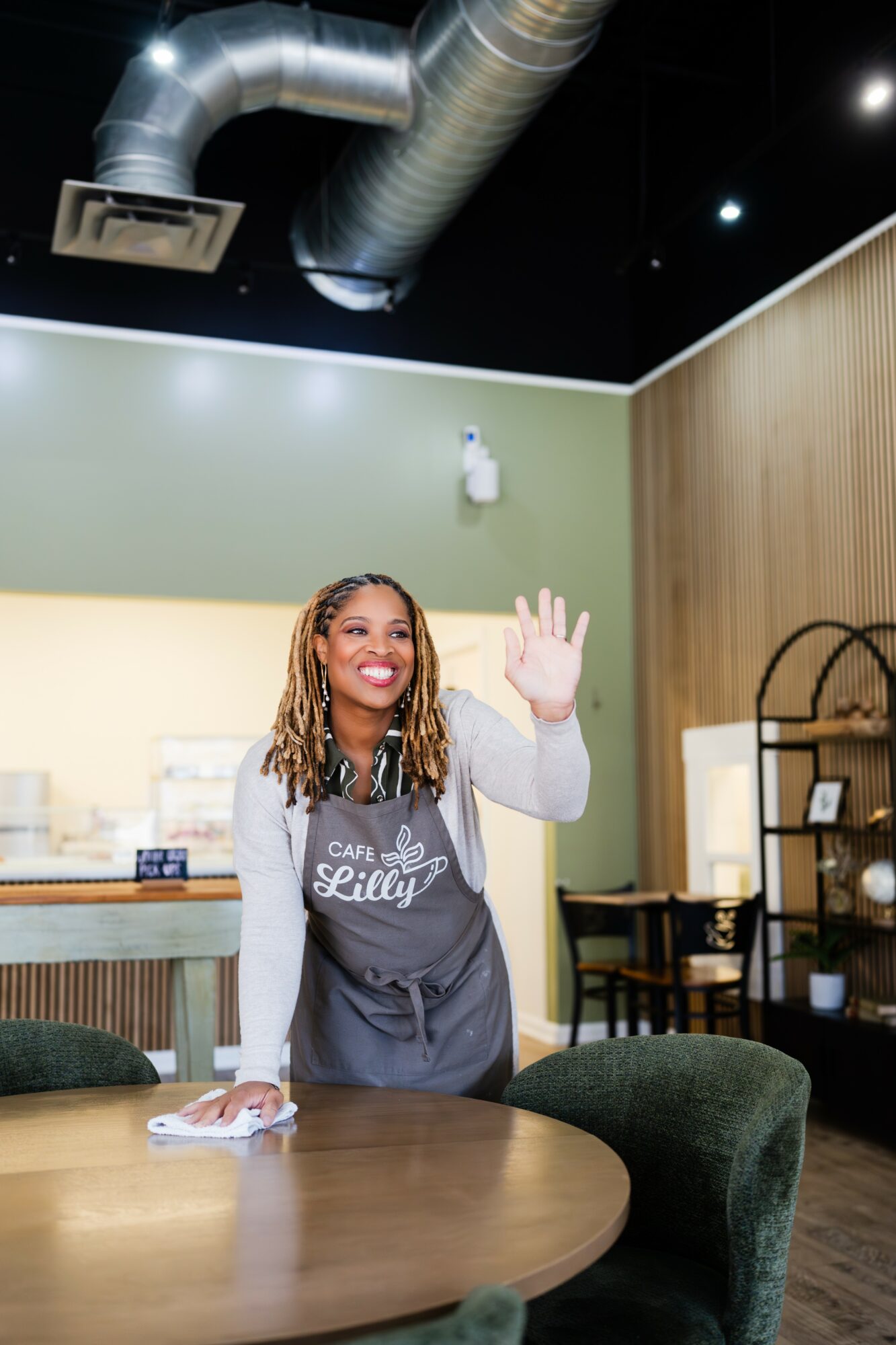 Smiling woman waving in a cafe, wearing an apron, with a table and shelves in the background.