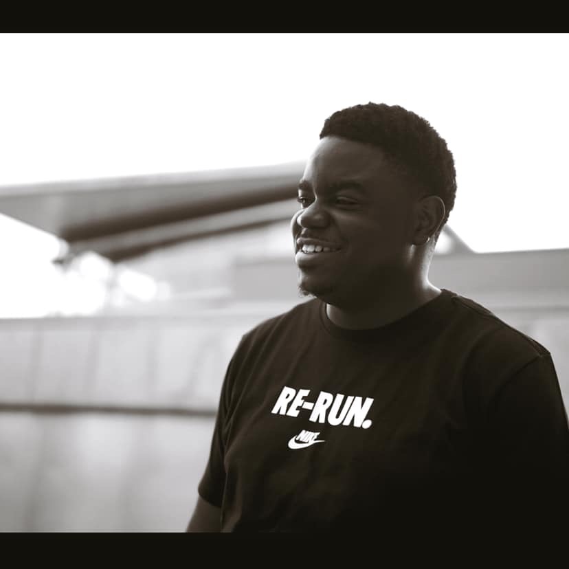 Man smiling outdoors wearing a black T-shirt with 'RE-RUN' and Nike logo, background blurred.
