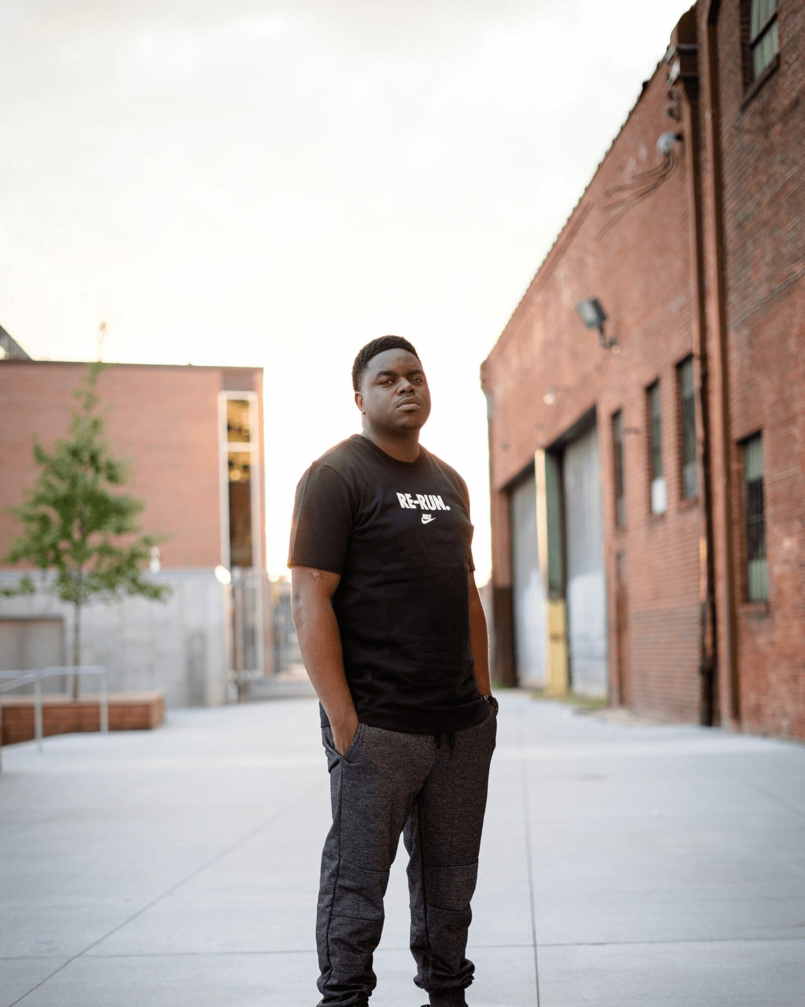 Young man standing outdoors in an urban area with brick buildings, wearing a black t-shirt and dark pants.