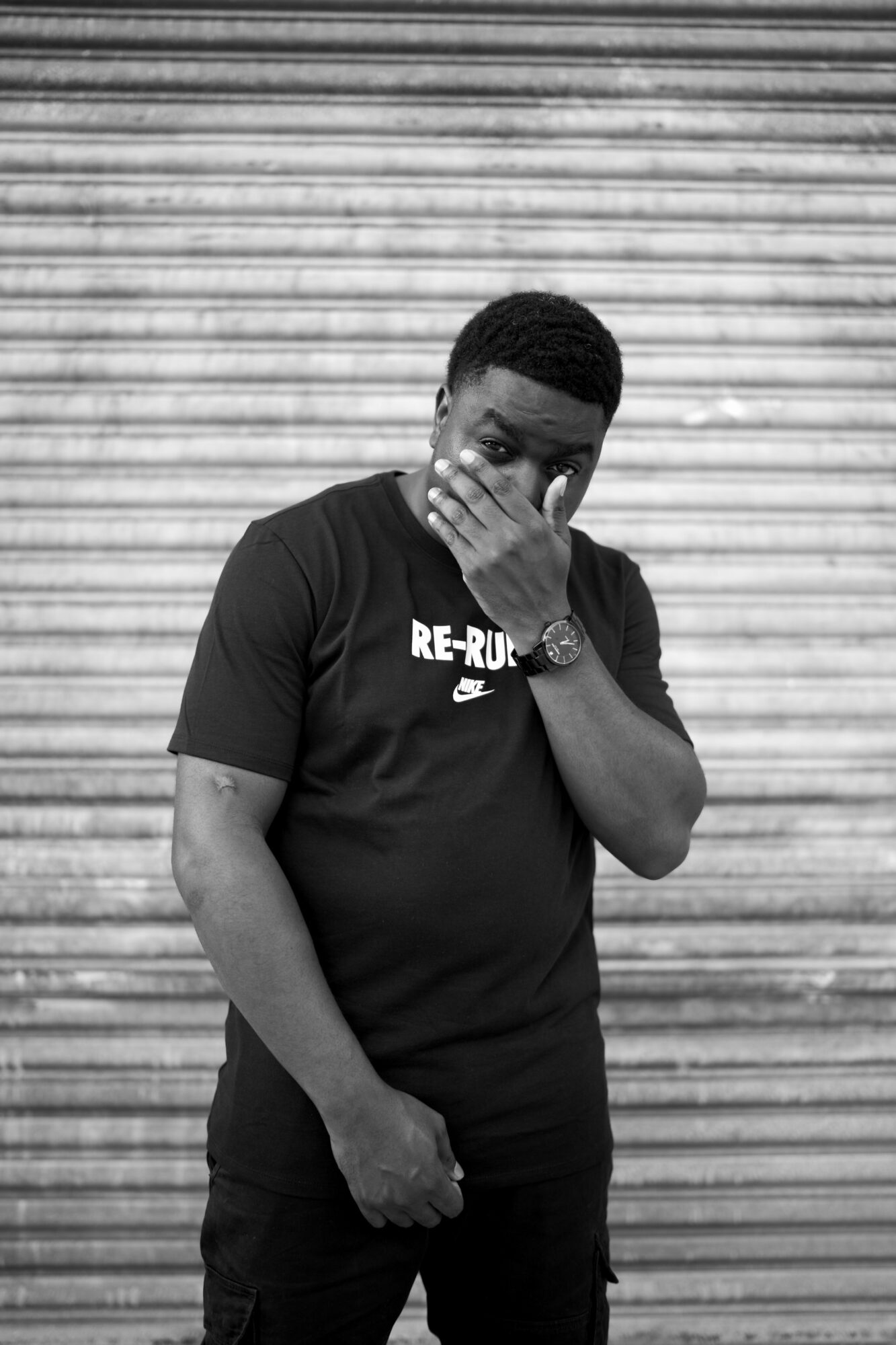 Young man covering his face with his hand, standing in front of a horizontal metal shutter, wearing a black T-shirt.