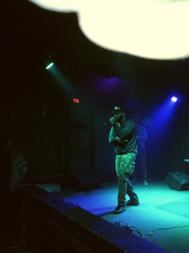 Performer on stage with microphone, illuminated by blue and purple lights, dark background, stage equipment visible.