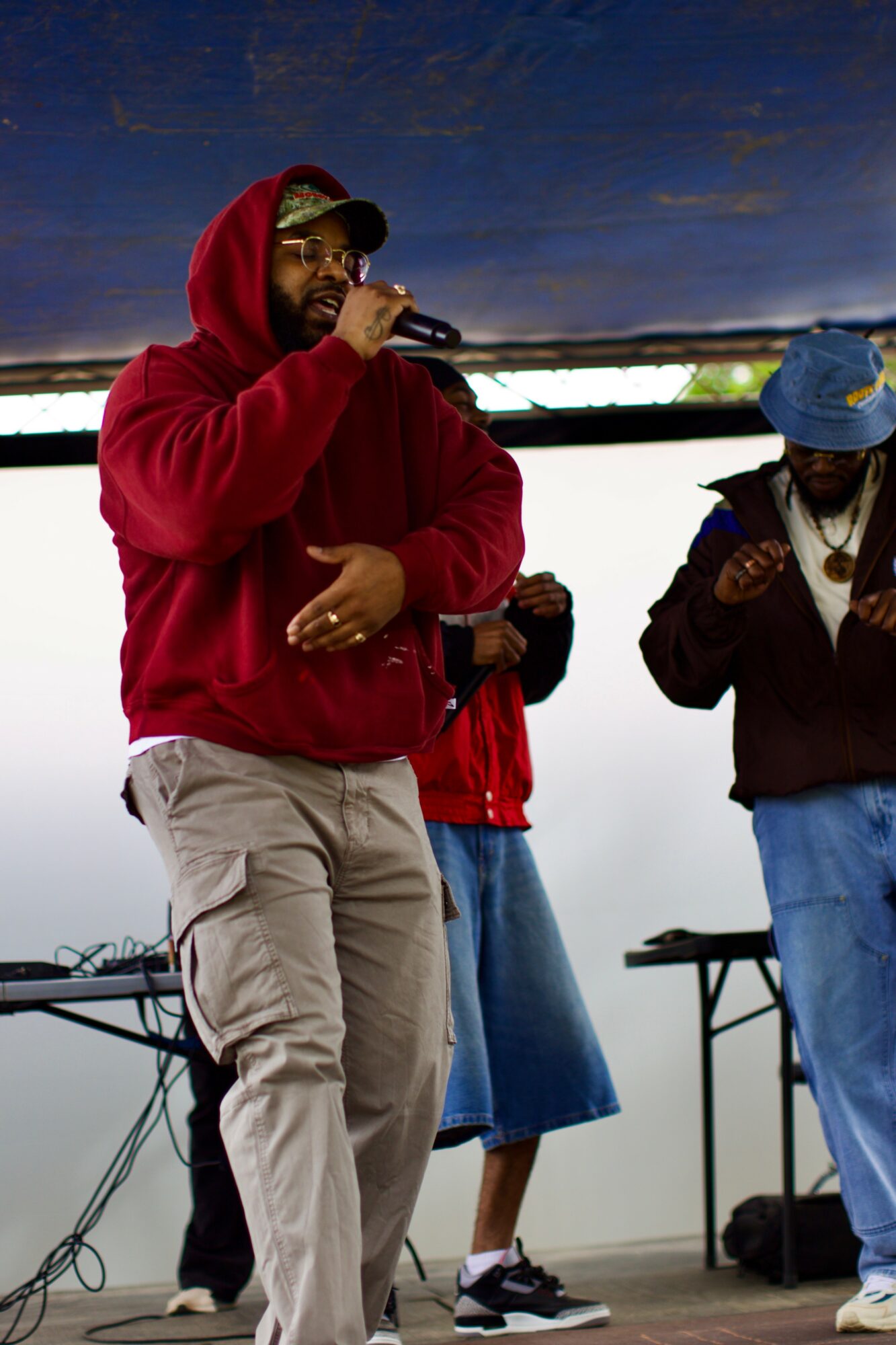 Person in red hoodie and glasses holding a microphone, with two other people on stage, outdoors, under a cloudy sky.
