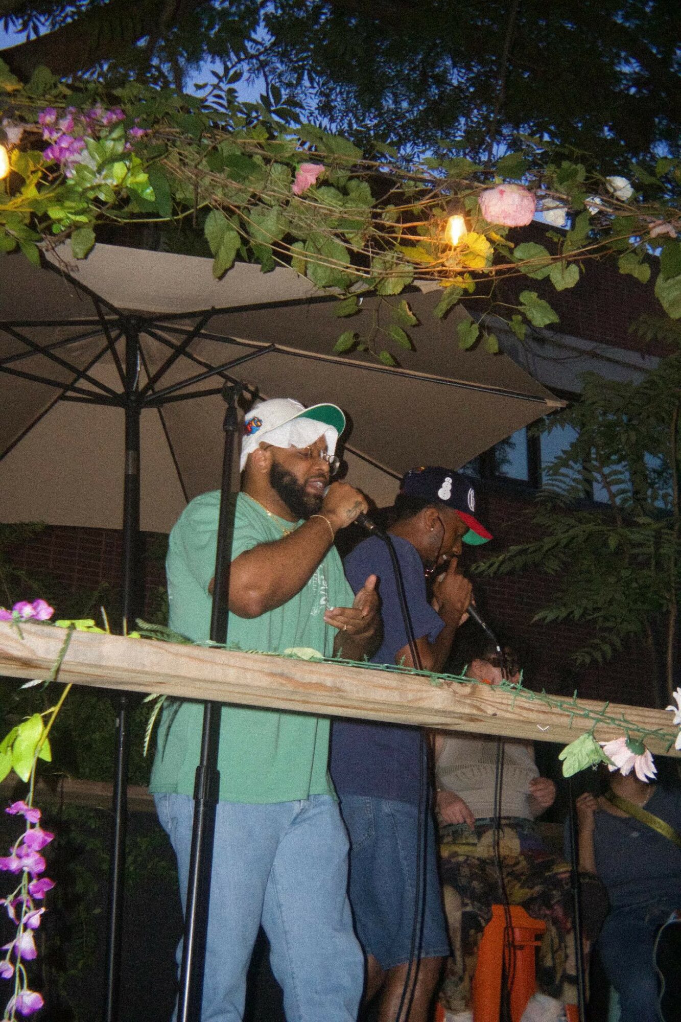 Two men are singing or speaking into microphones outdoors at night, with trees and string lights overhead.