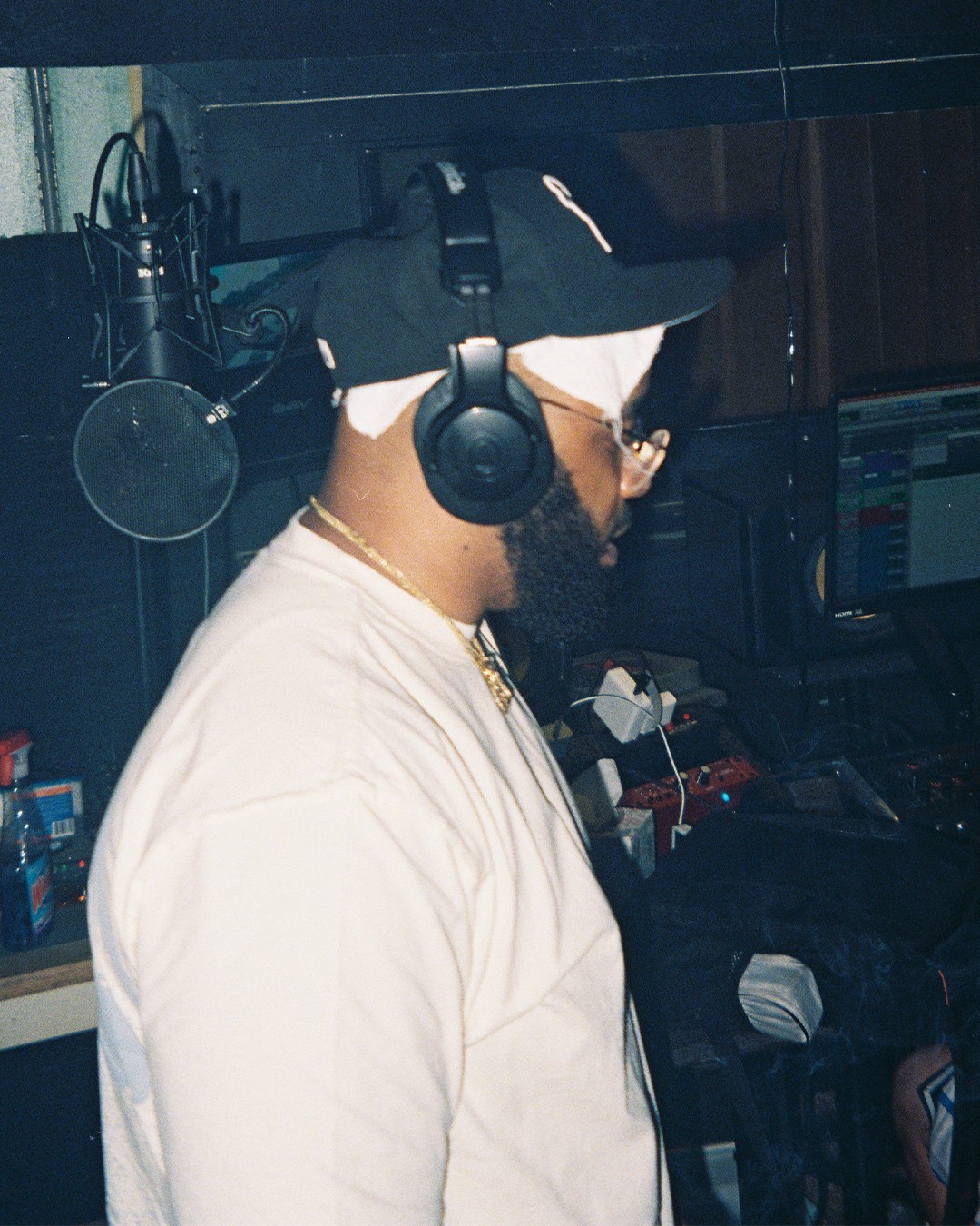 Person wearing headphones, cap, and glasses working at a computer in a recording studio.