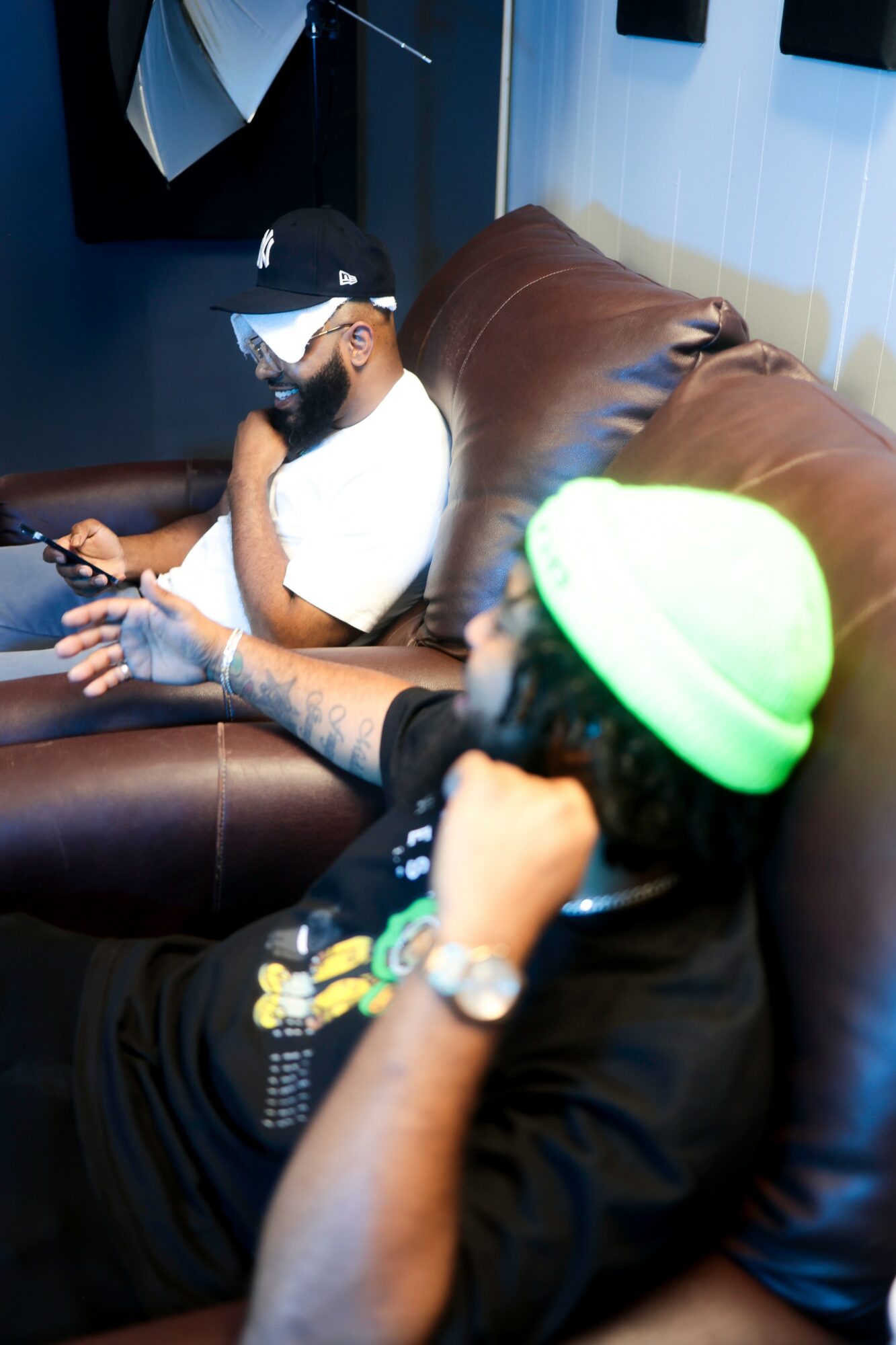 Two men sitting on a brown leather couch, one using a smartphone, the other wearing a green cap, in a room with dark walls.