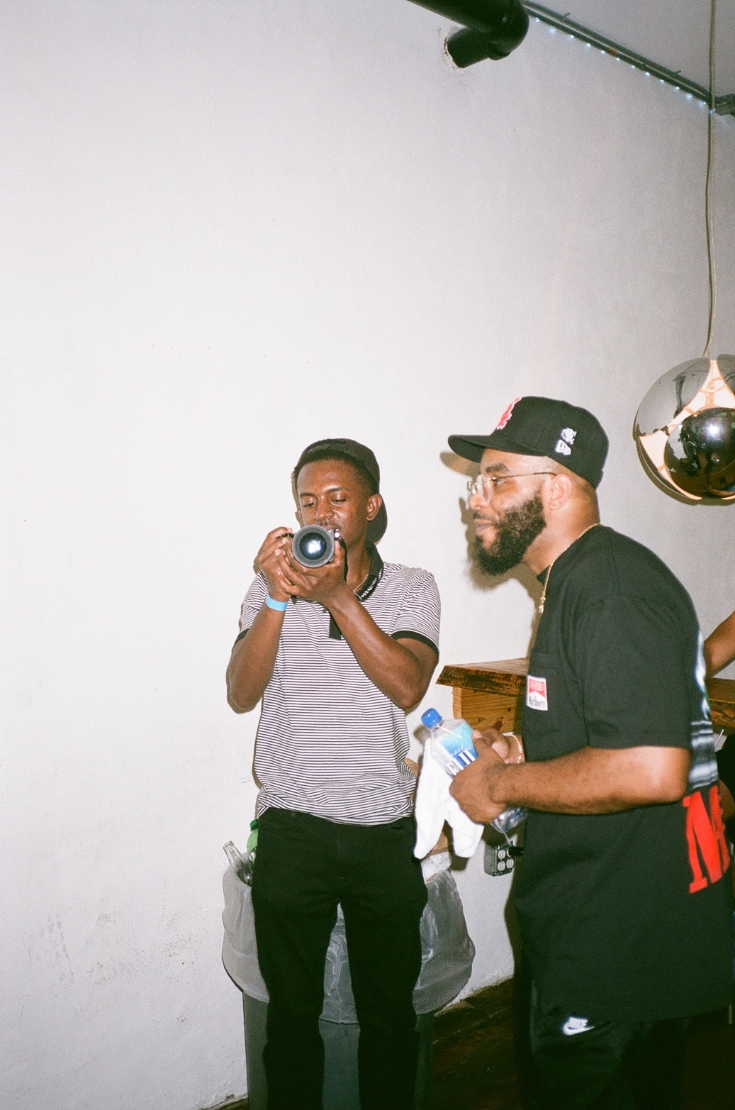 Two men standing indoors, one holding a camera and taking a photo, the other holding a water bottle, with a white wall background.