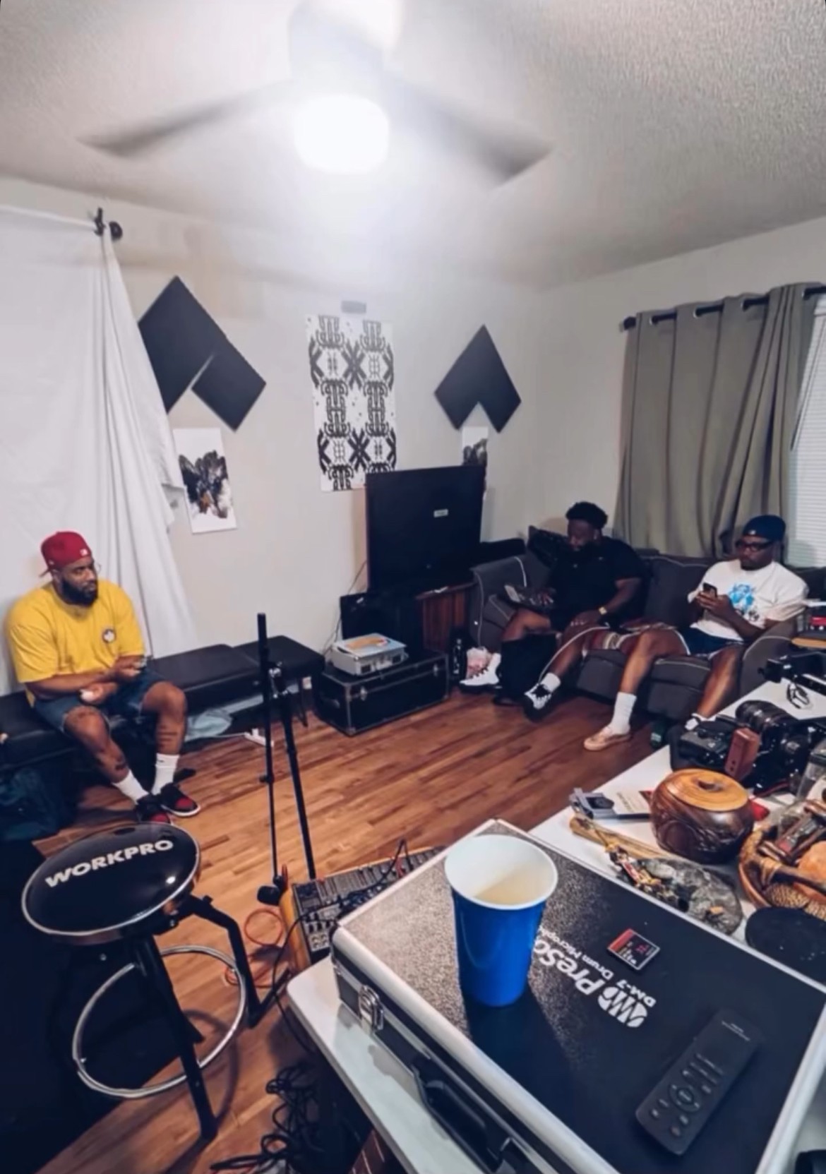Four people sitting in a living room with musical equipment, a TV, and curtains, with a wooden floor and ceiling fan.