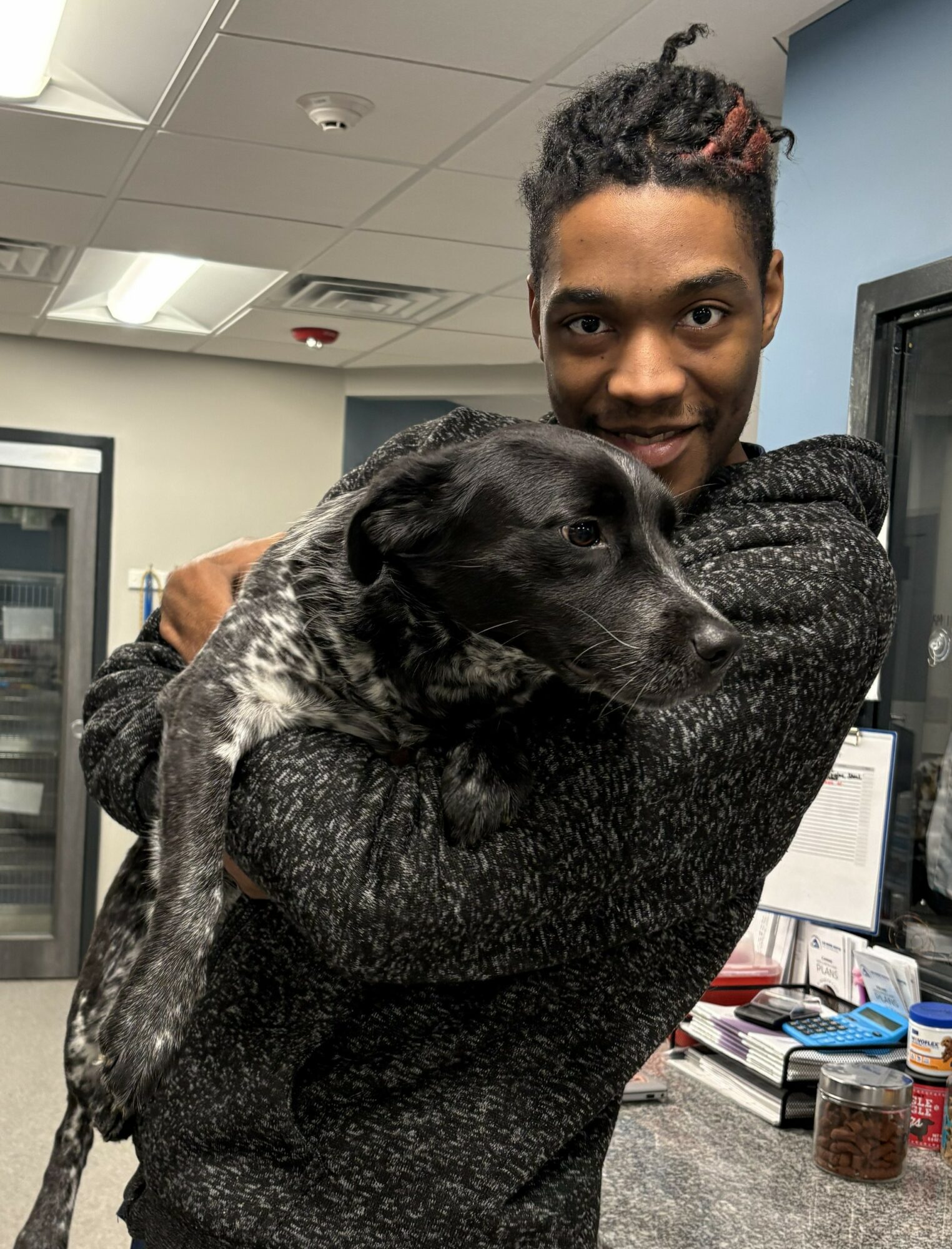 Person holding a black and white dog indoors, smiling at the camera, with computer monitors and office supplies in the background.