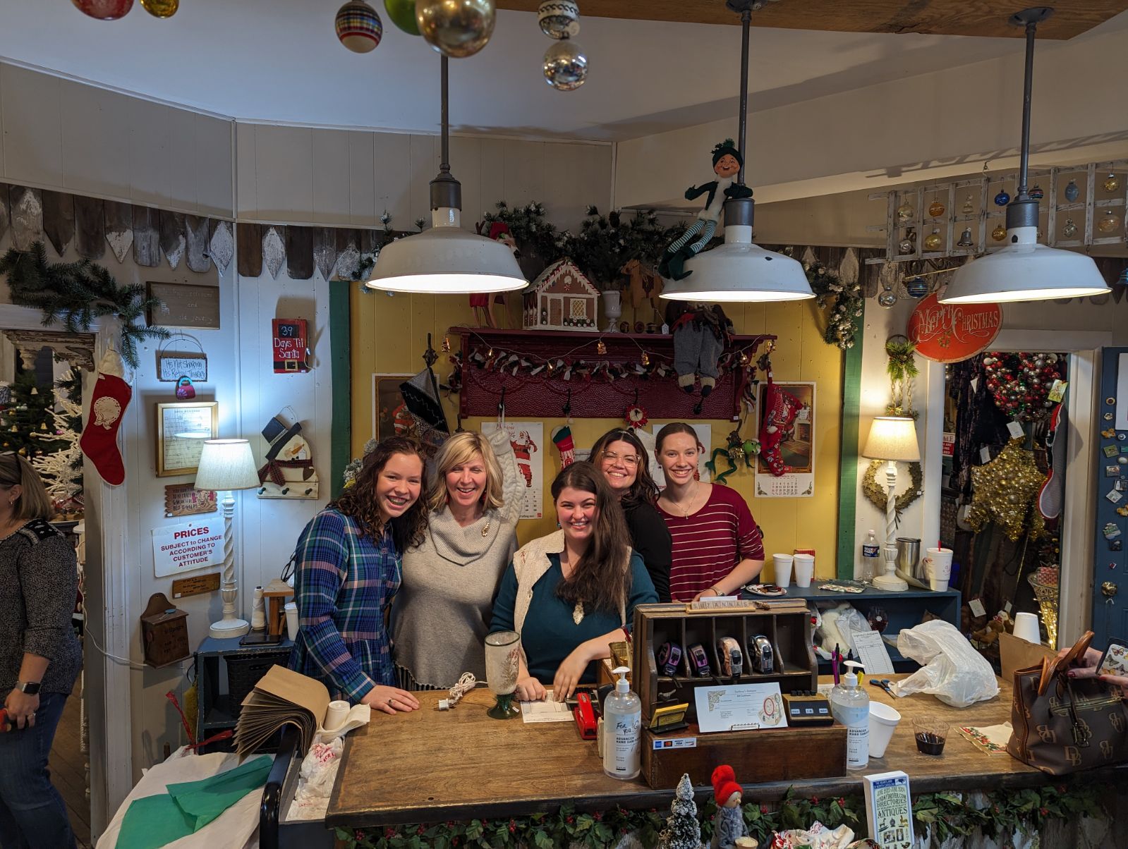 Five people stand behind a counter in a decorated room with holiday decorations and warm lighting.