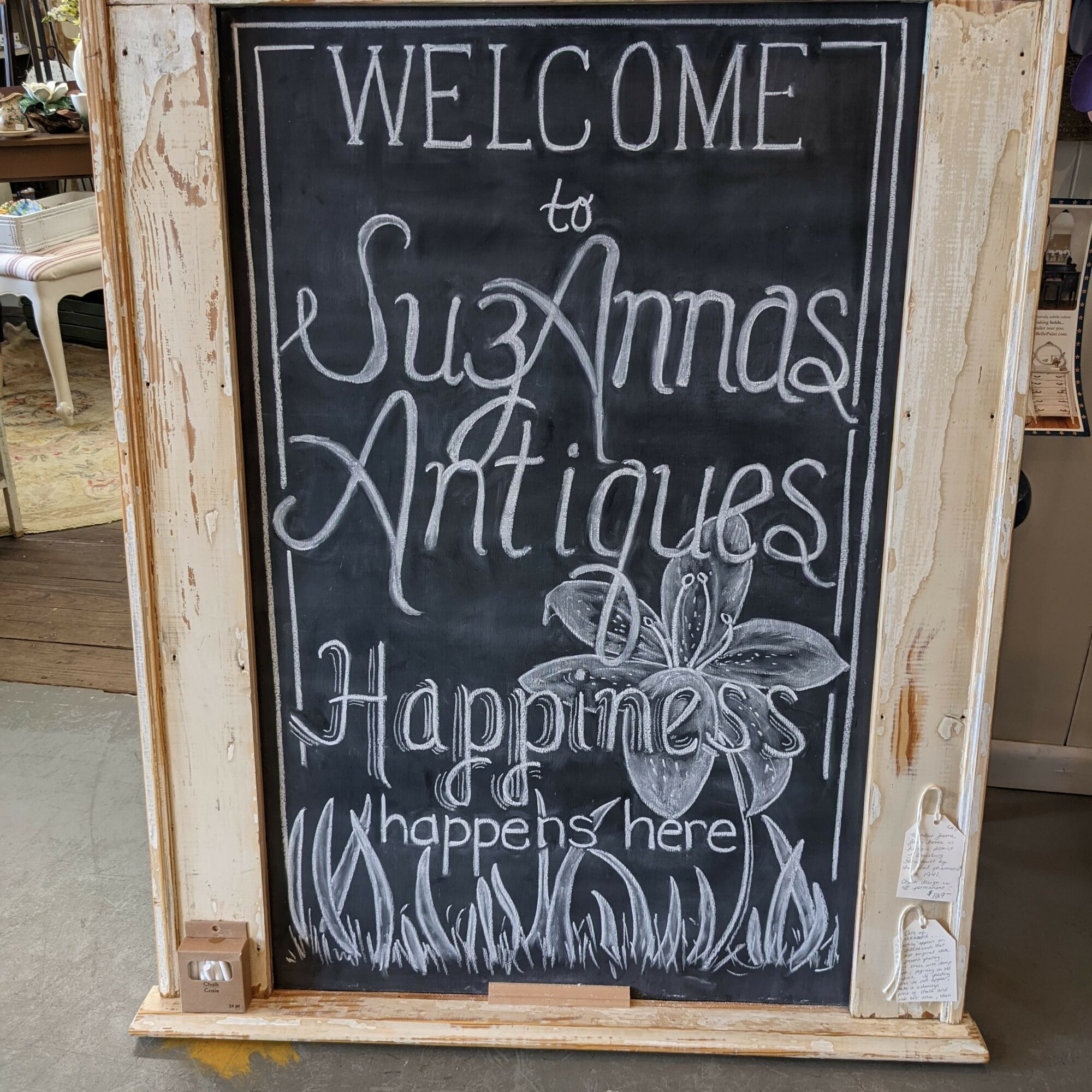 Blackboard sign with white chalk writing welcoming to SuzAnna's Antiques, Happiness happens here, with a flower drawing and grass at the bottom.