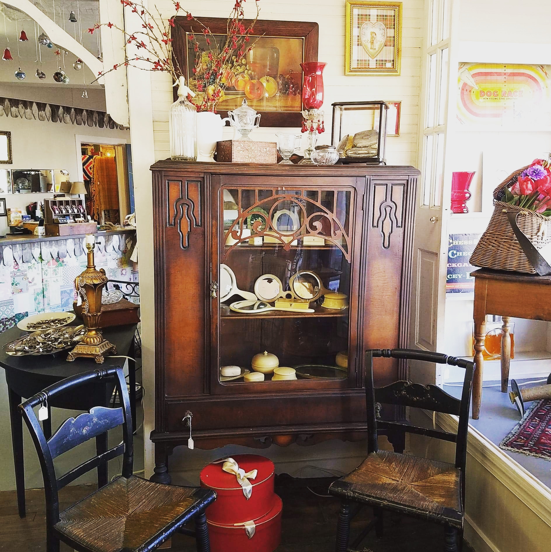 Wooden display cabinet with glass doors, decorated with vases, framed pictures, and various dishes inside, in a room with other furniture.