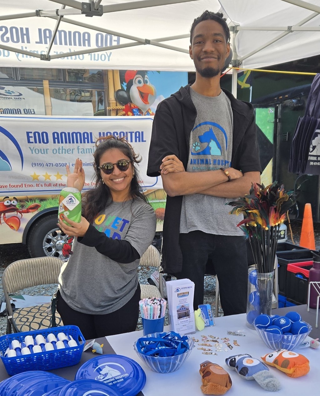 Two people standing behind a table at an outdoor event, smiling, with a banner and animal-themed decorations.