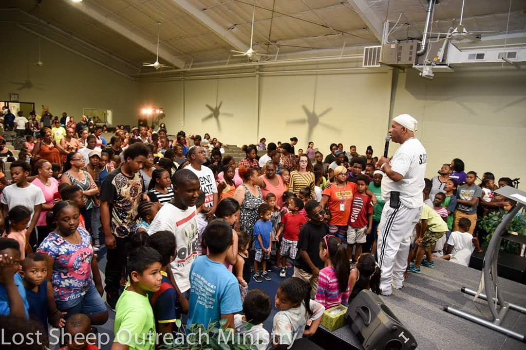 Large group of children and adults gathered indoors, with a speaker on stage addressing the crowd, shadows on the wall.