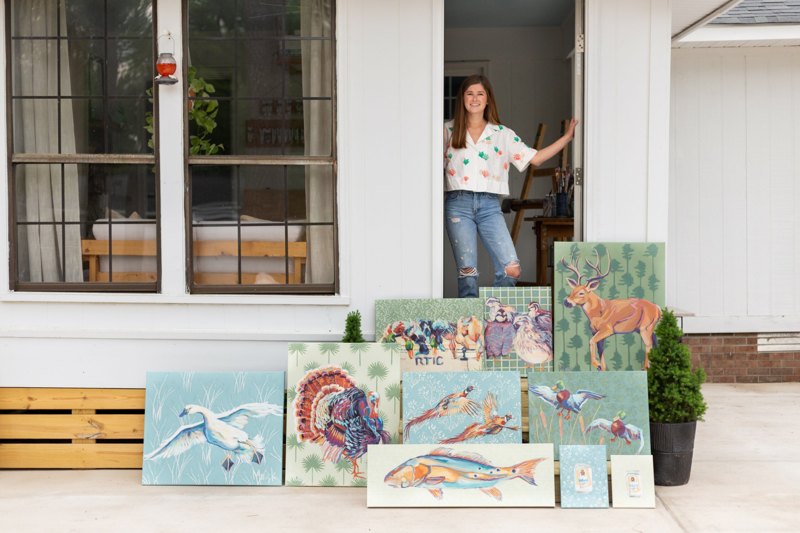 Woman standing in doorway with paintings of animals outside, potted plant nearby, wooden bench, and window with curtains behind.