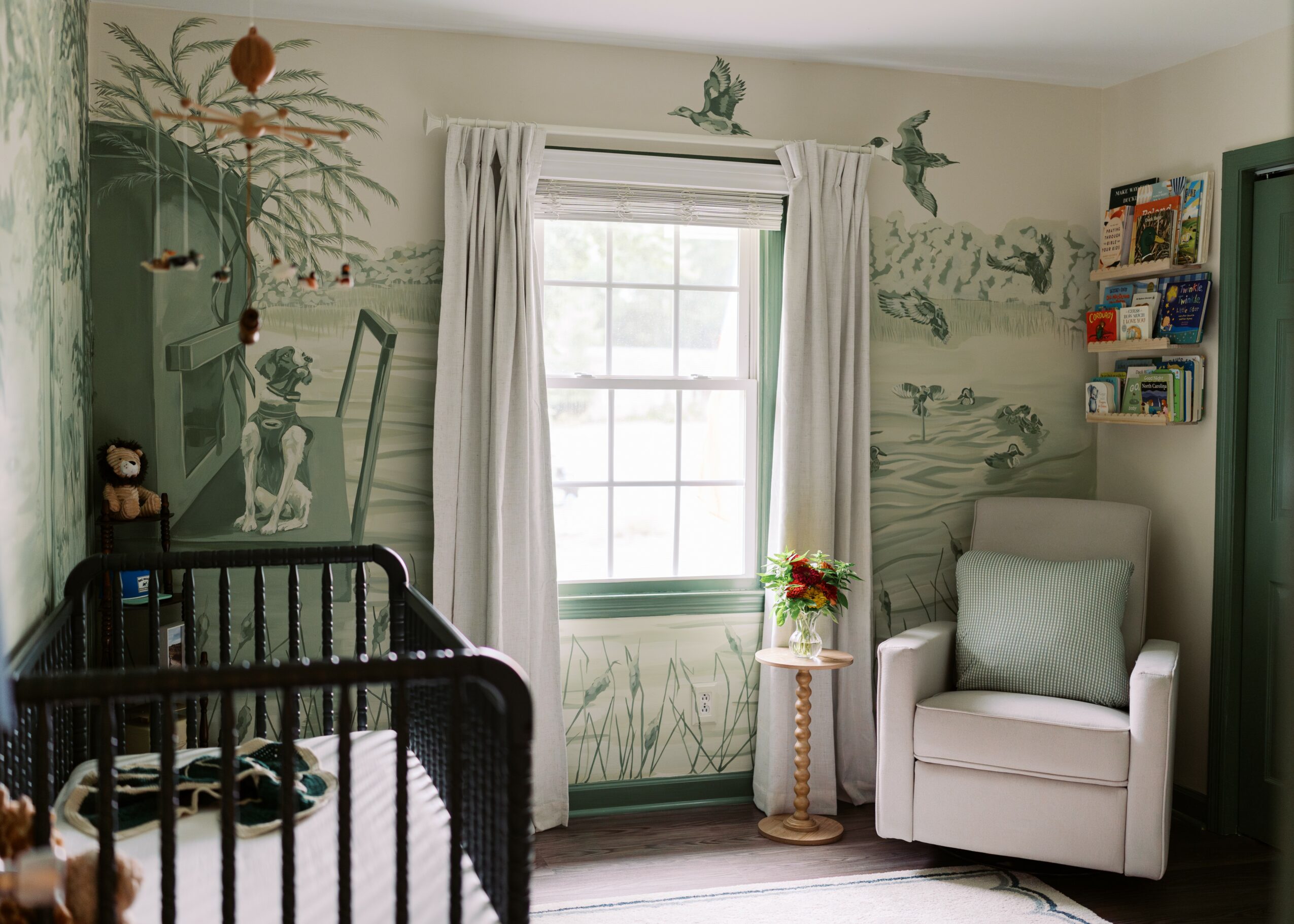 Room with a window, white curtains, armchair, small table with flowers, and mural of nature scenes on walls.