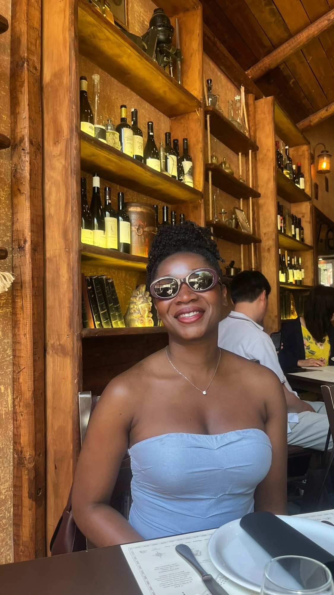 Smiling woman with glasses in a strapless top sitting at a table in a wooden bar or restaurant, shelves with bottles behind her.