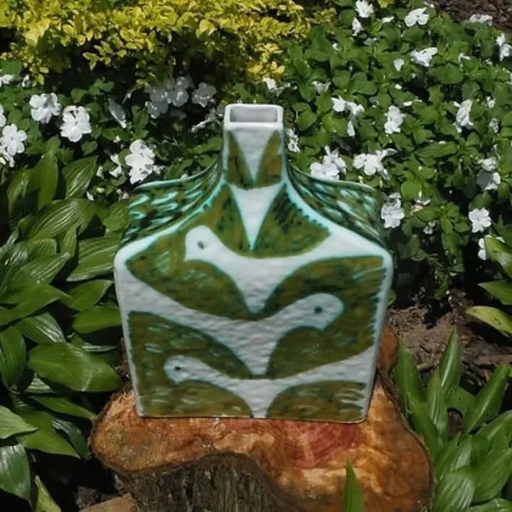 Decorative ceramic vase with green and white pattern, placed on a tree stump among green plants and white flowers.