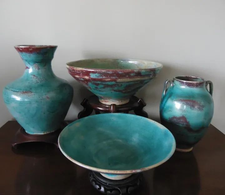 Four ceramic bowls and vases with turquoise, red, and green glaze on dark wooden stands, arranged on a dark surface.