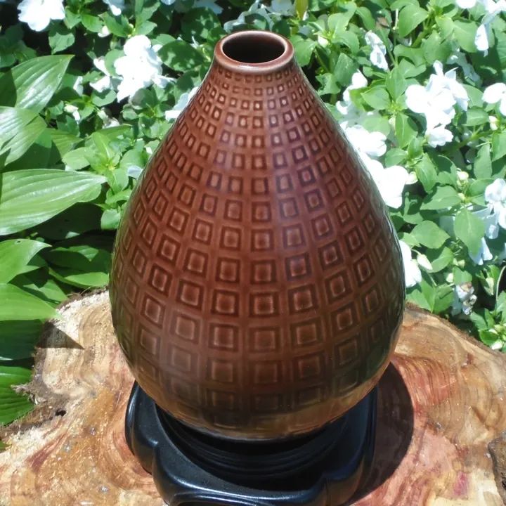 Brown vase with geometric pattern on a wooden surface, surrounded by green leaves and white flowers.