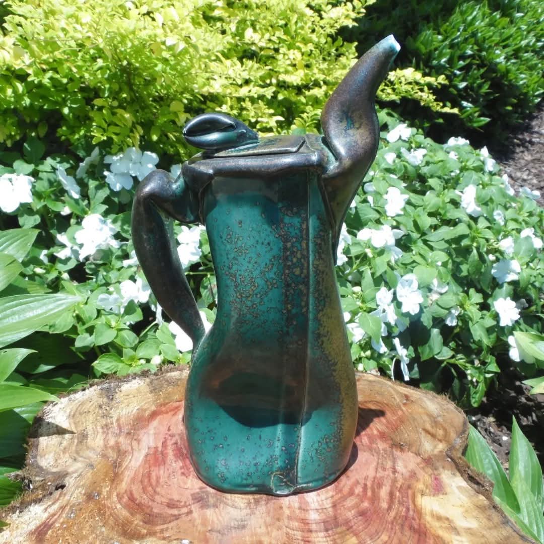 A metal sculpture of a stylized female figure with an elongated body and abstract features, placed outdoors among green plants and white flowers.