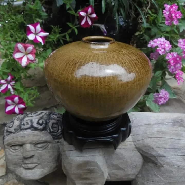 Round ceramic vase with a small opening, placed on a black stand among pink and purple flowers and stone sculptures.