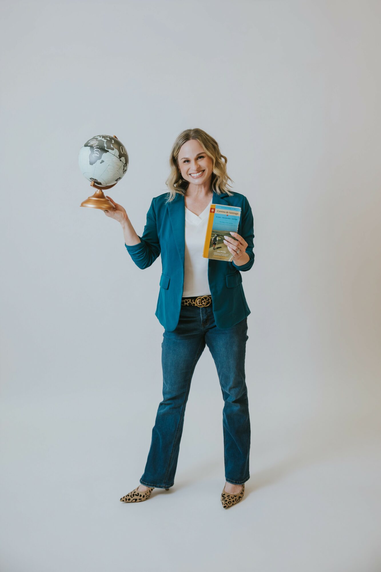 Woman holding a globe in one hand and a book in the other, smiling, standing against a plain background.