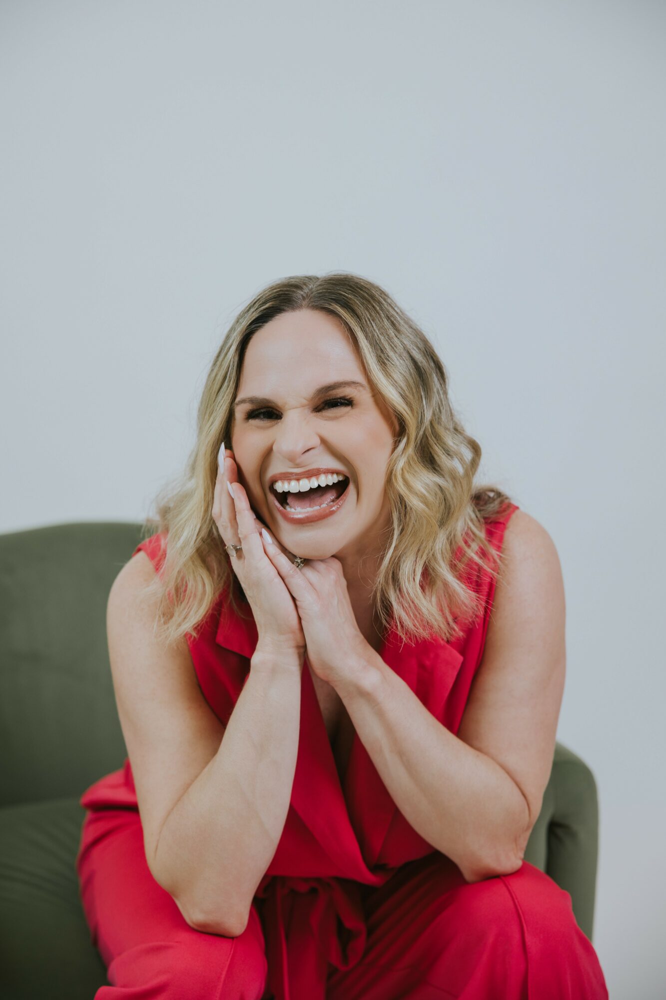 Woman with blonde hair laughing, hands on face, wearing a red dress, sitting on a green chair against a plain background.