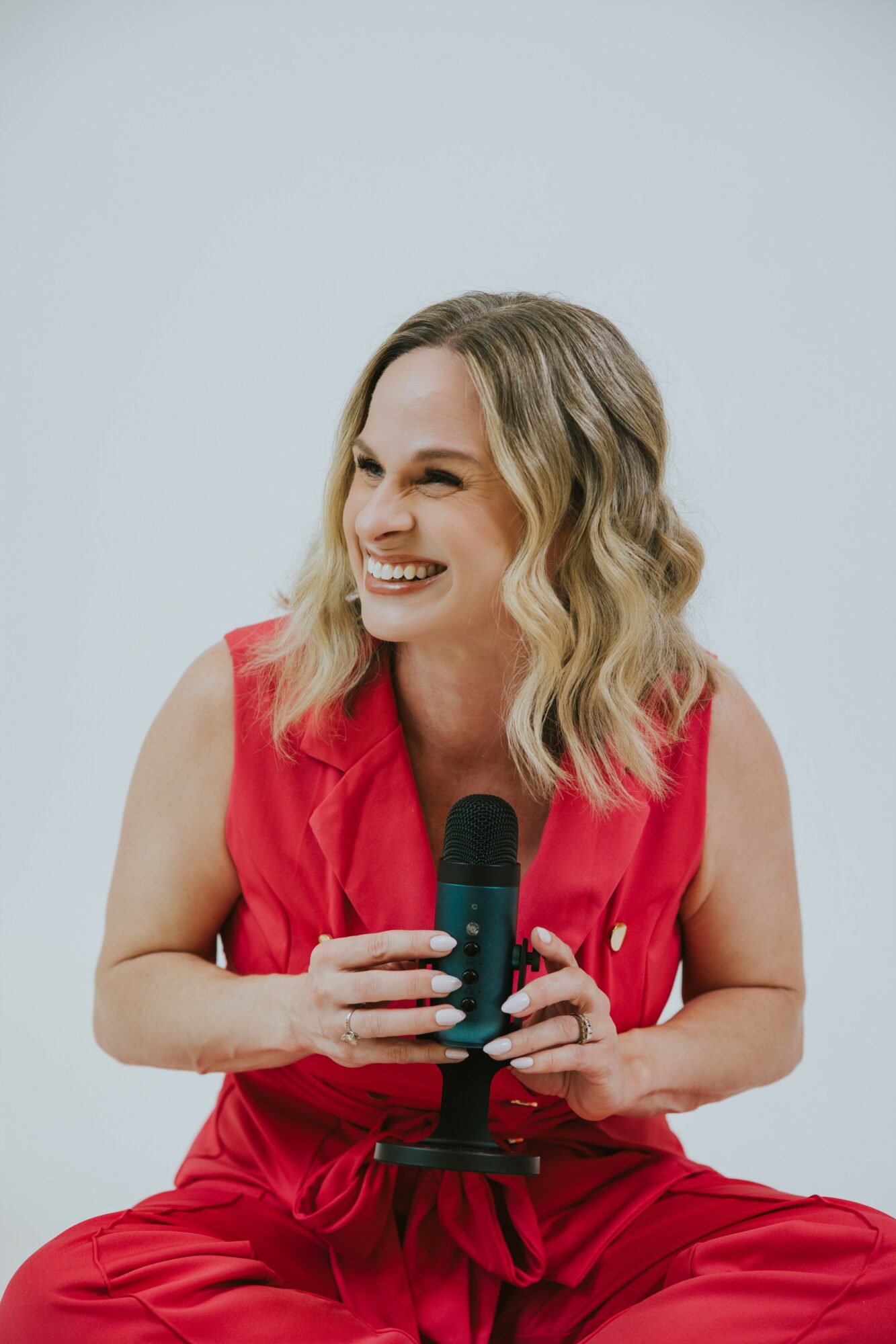 Woman with wavy blonde hair smiling, holding a microphone, wearing a sleeveless red outfit, against a plain light background.