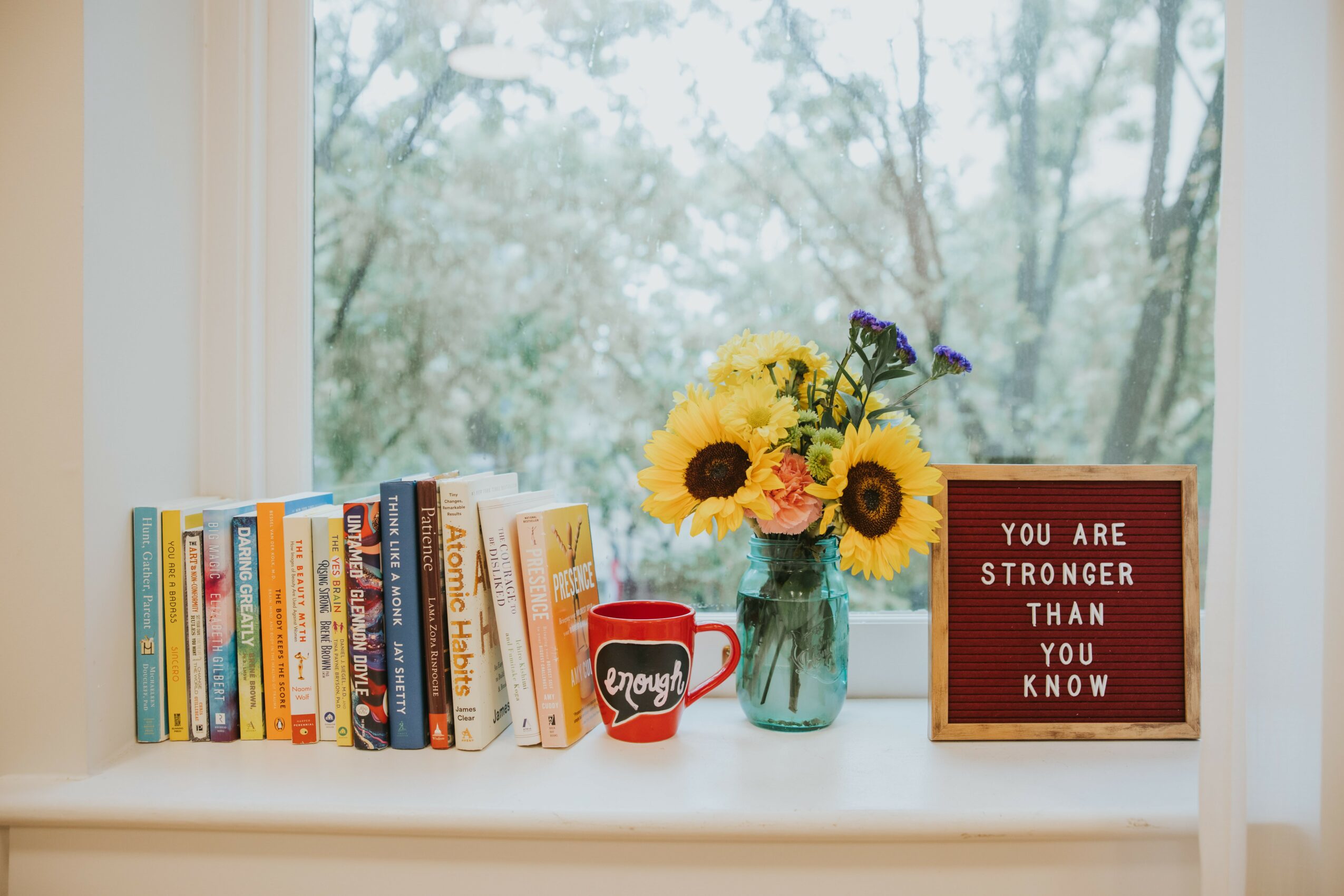Books, a red mug, and a vase with sunflowers and purple flowers on a windowsill, with trees outside.