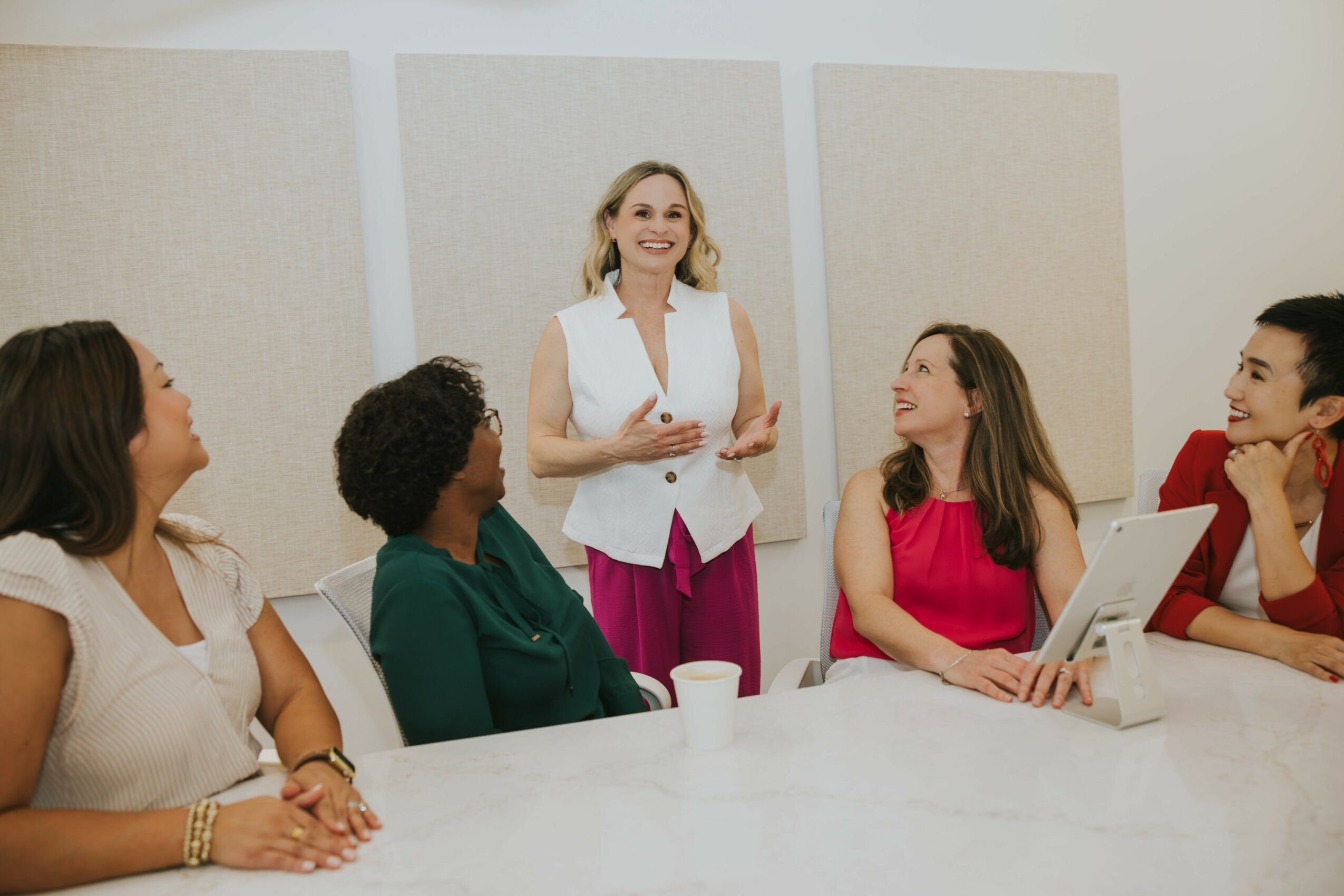 A woman stands and speaks to a group of four women seated at a table in a meeting room.