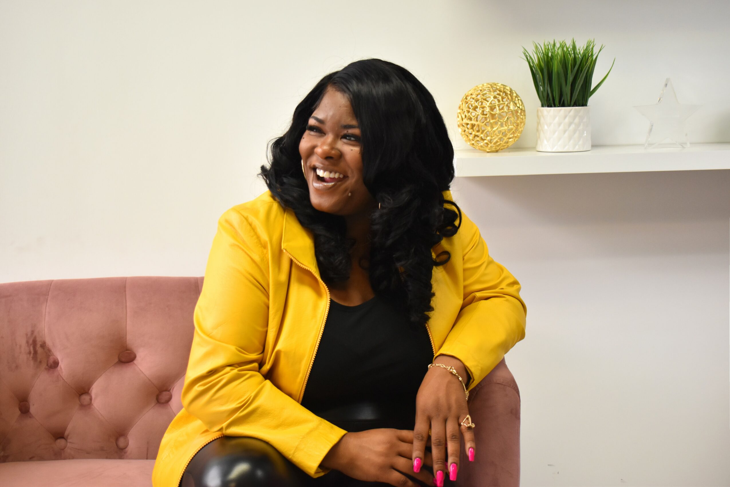 Woman with black hair smiling, wearing a yellow jacket and black top, sitting on a pink sofa, with a white shelf and plants behind her.
