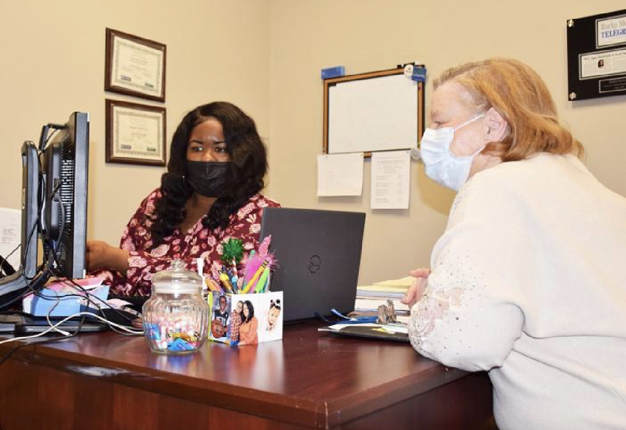 Two women wearing masks sit at a desk with a computer, papers, and office supplies in a professional setting.