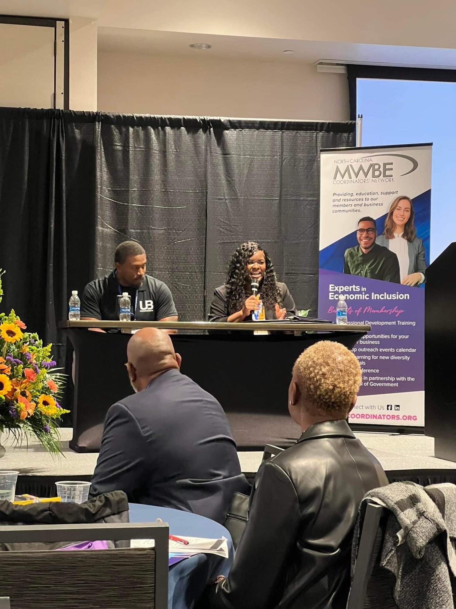 Three people seated at a panel table on stage, with a black curtain backdrop and a MWBE banner, audience in foreground.