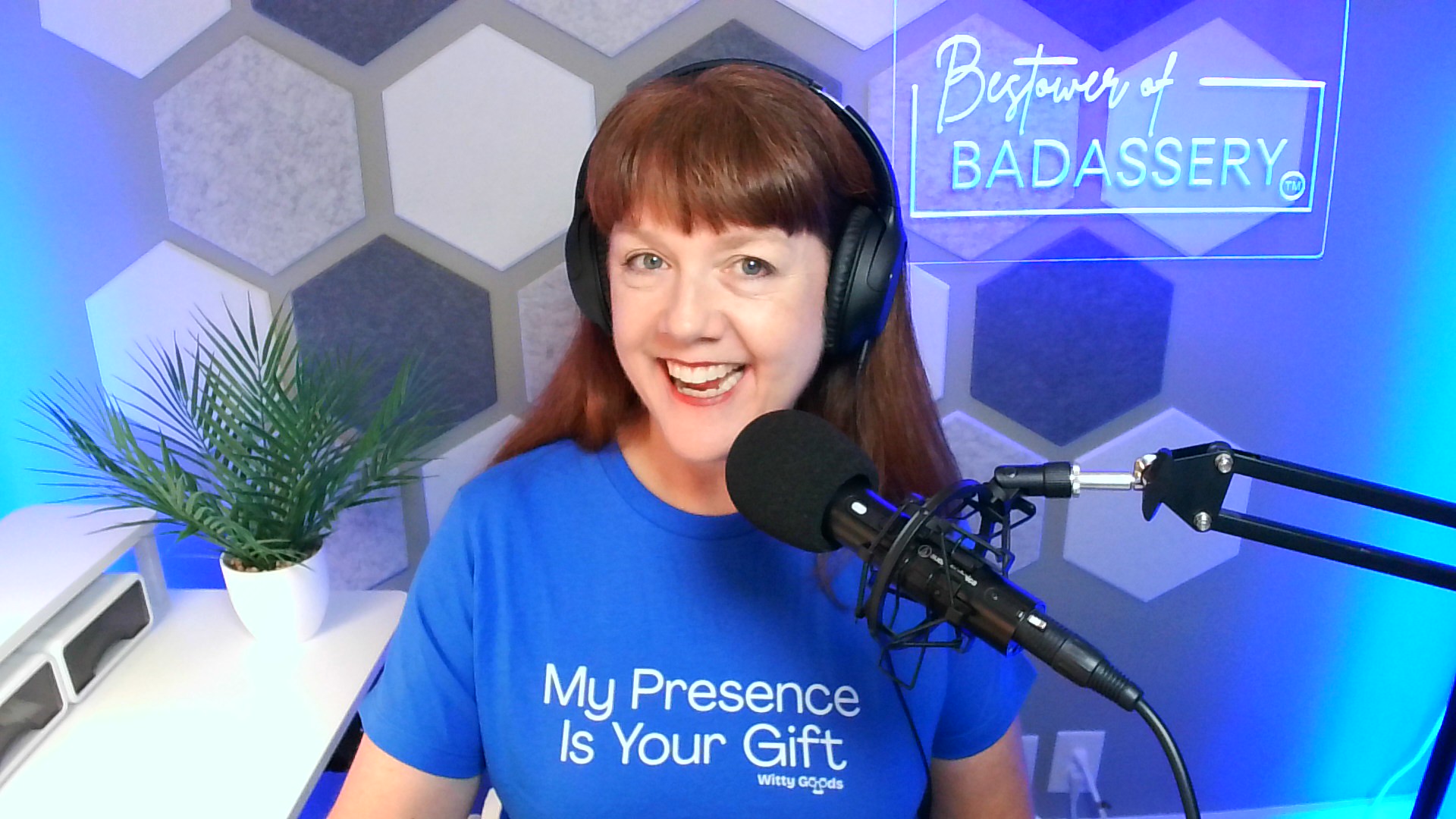 Woman with red hair wearing headphones, smiling at microphone in a studio with hexagonal wall pattern and a plant.