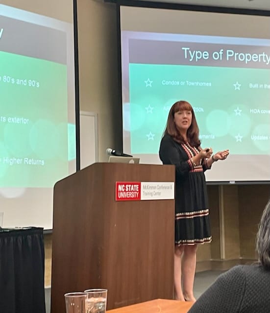 Woman giving a presentation at a podium in a classroom or conference room, with slides projected behind her.