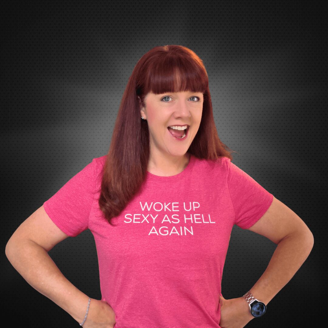 Woman with red hair smiling, wearing a pink T-shirt with text, against a dark background.