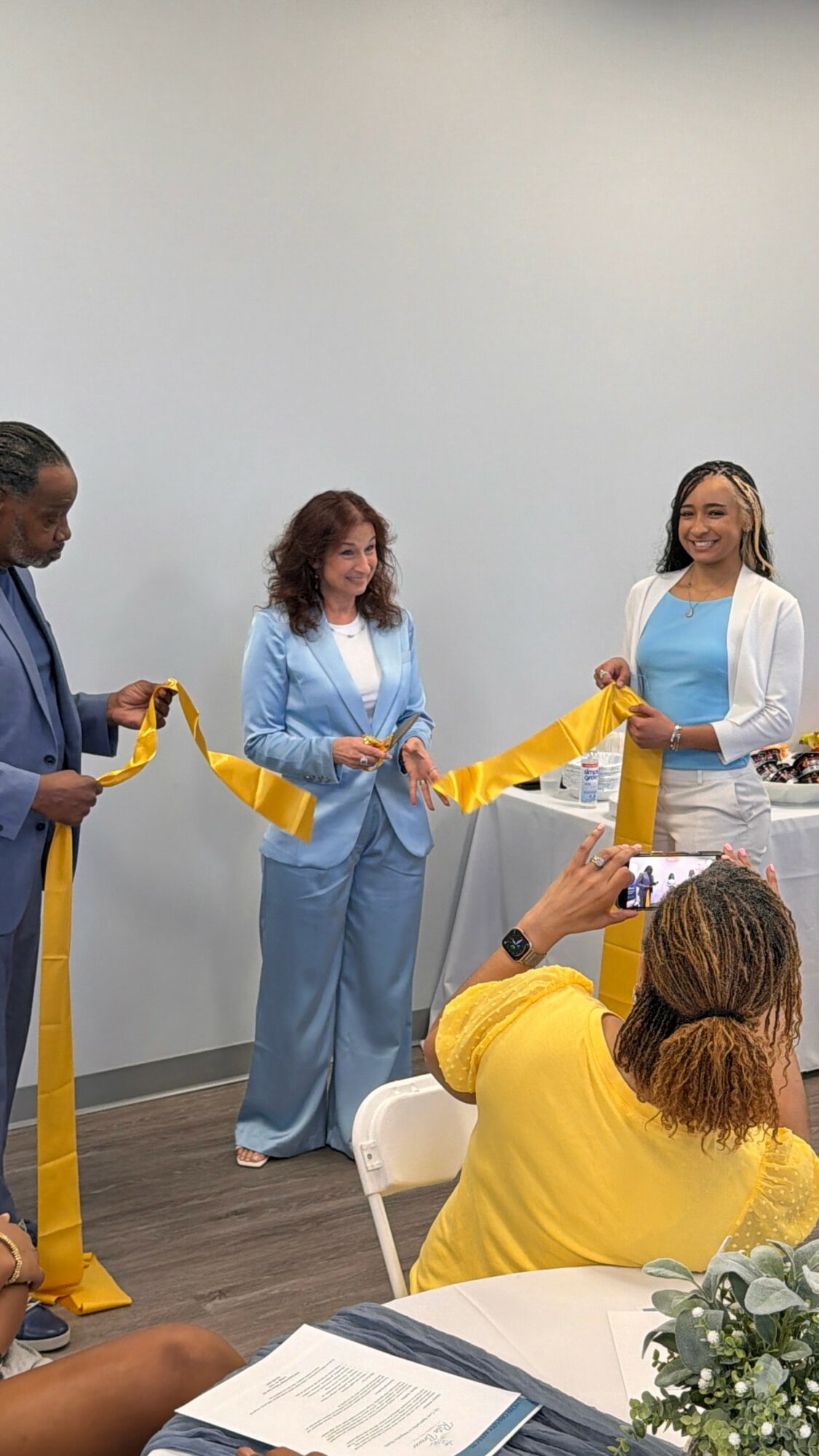 Three women stand holding a yellow ribbon, with two people seated in front taking photos, in a room with a plain wall.